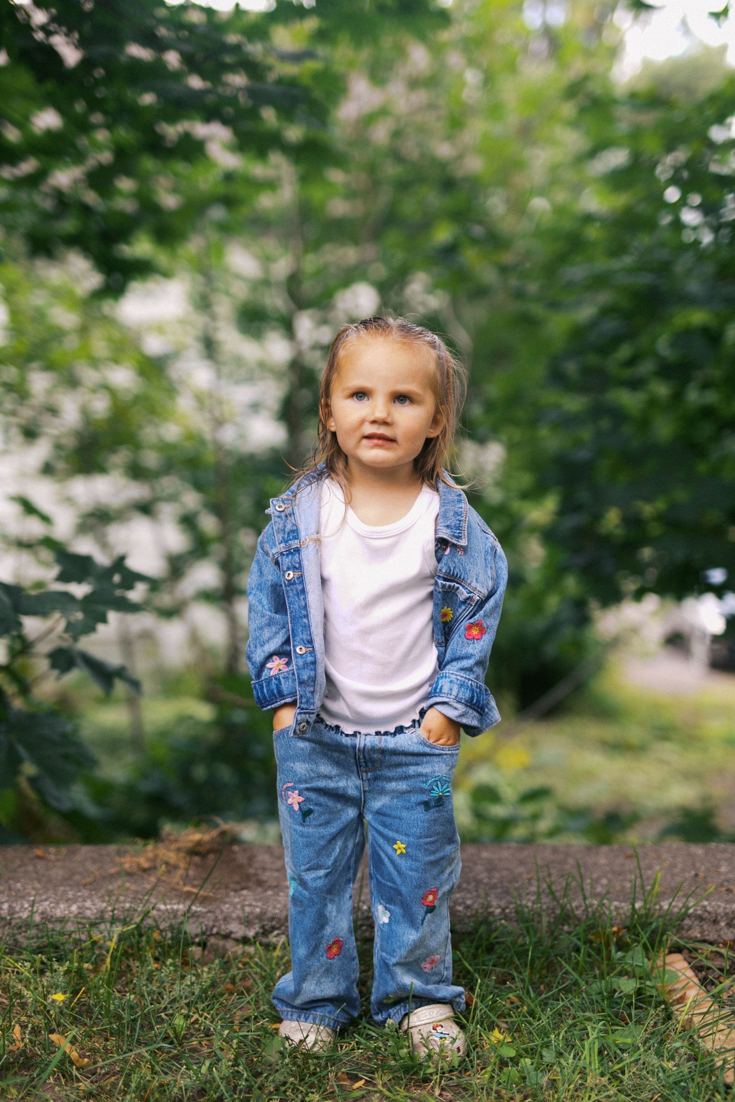 Little girl in floral embroidered denim jacket and jeans standing outdoors in green garden.