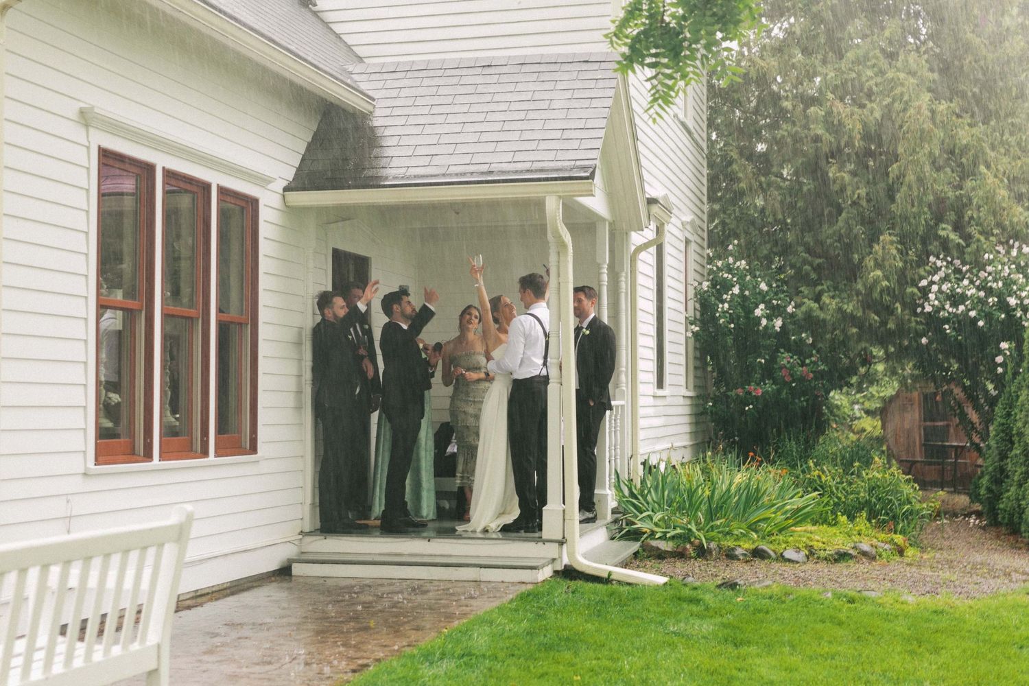 A view of a white country house with a porch where a bride and friends dance while it rains during a wedding.