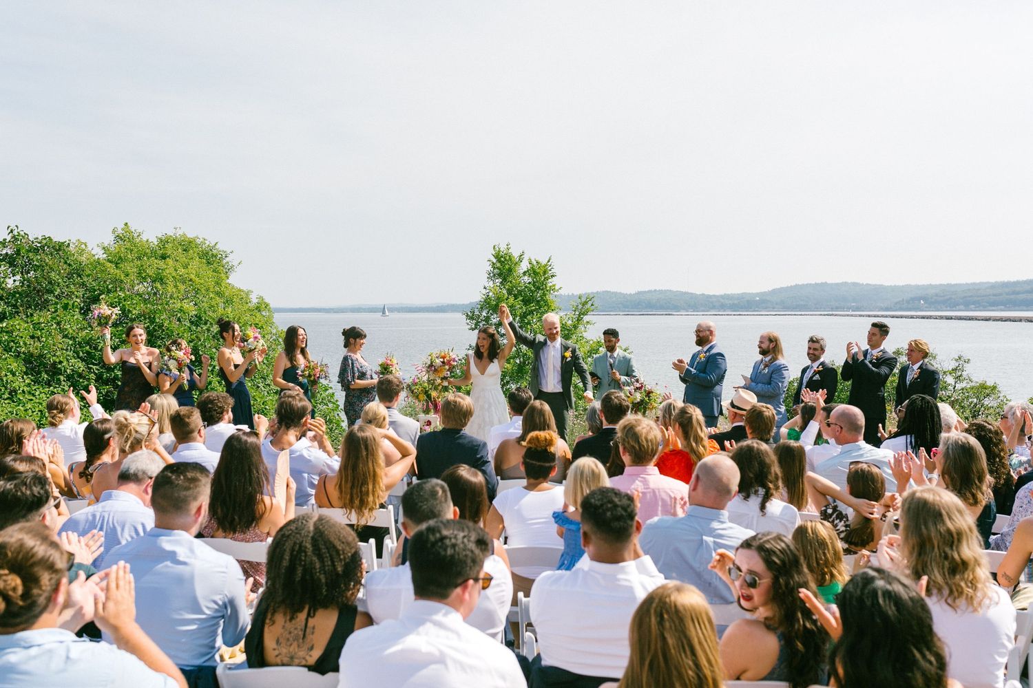 Wedding guests seated in white chairs for an outdoor ceremony overlooking the water.