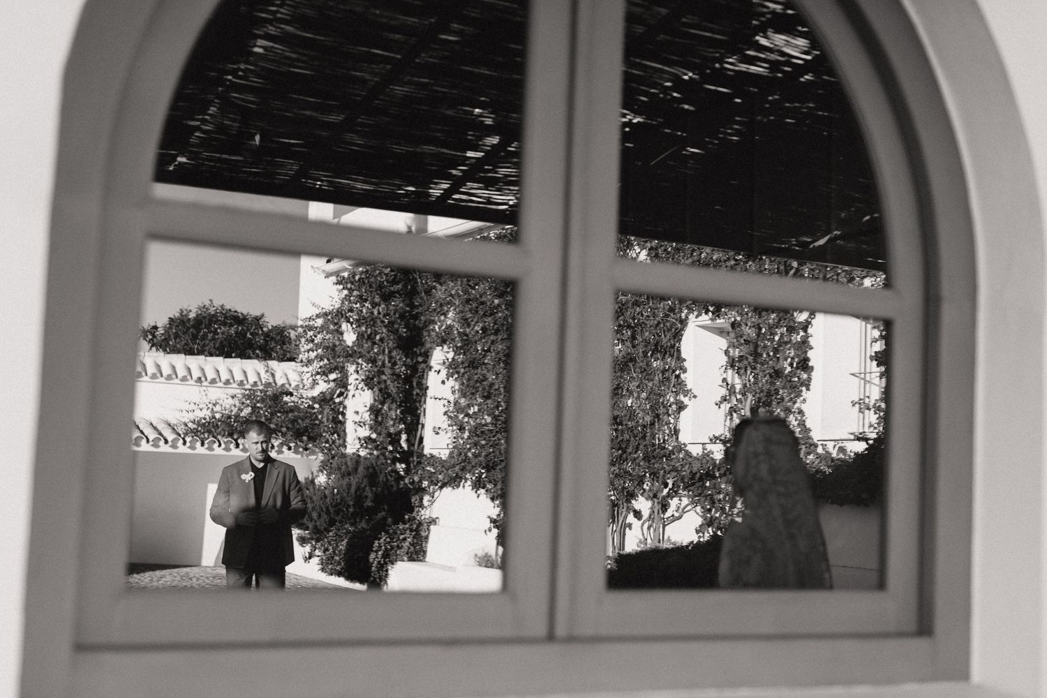 Black and white artistic reflection of wedding couple captured through arched window with garden backdrop.
