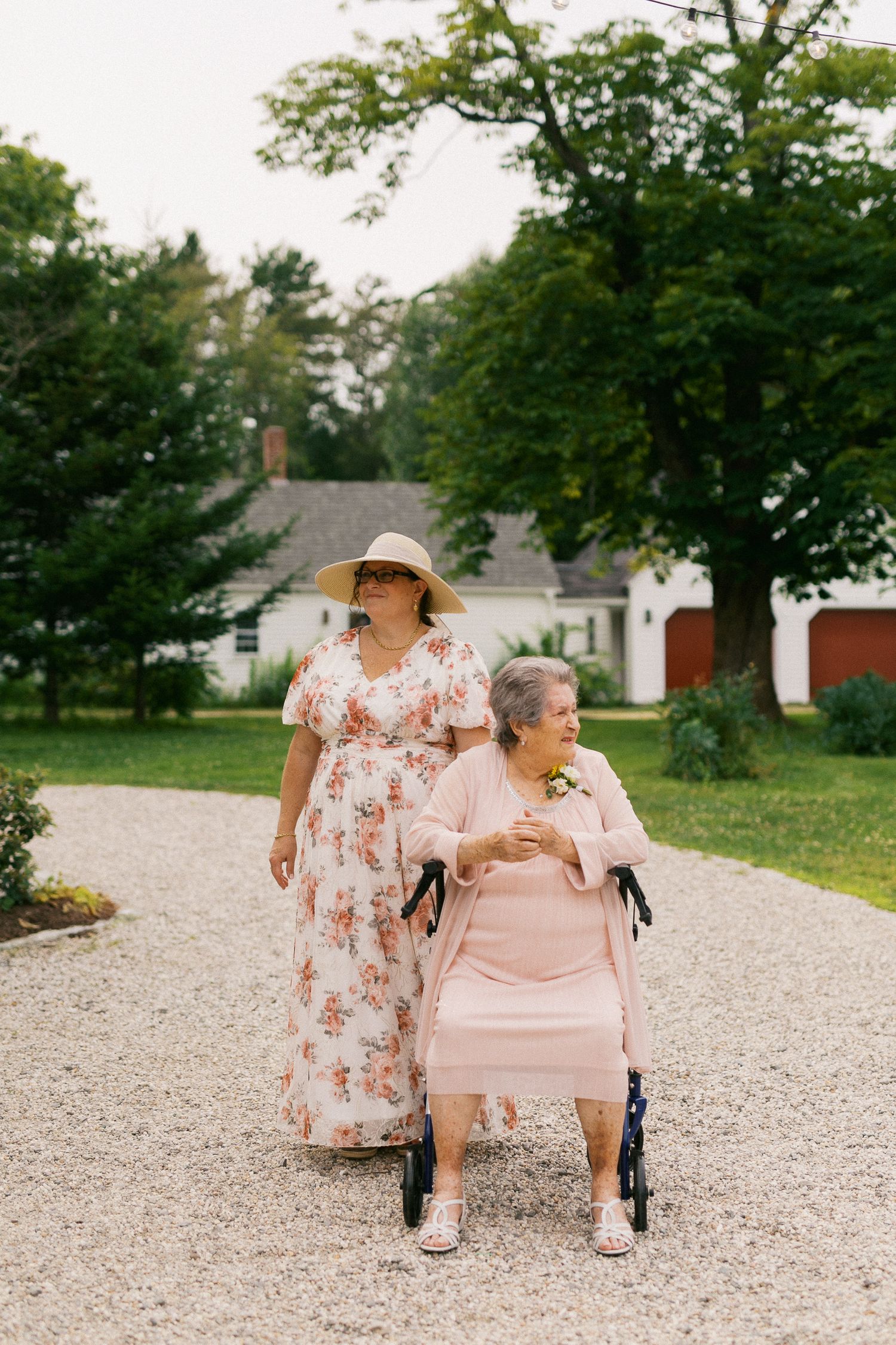Elderly woman in pink dress using a walker on gravel path, accompanied by woman in floral dress and sun hat.