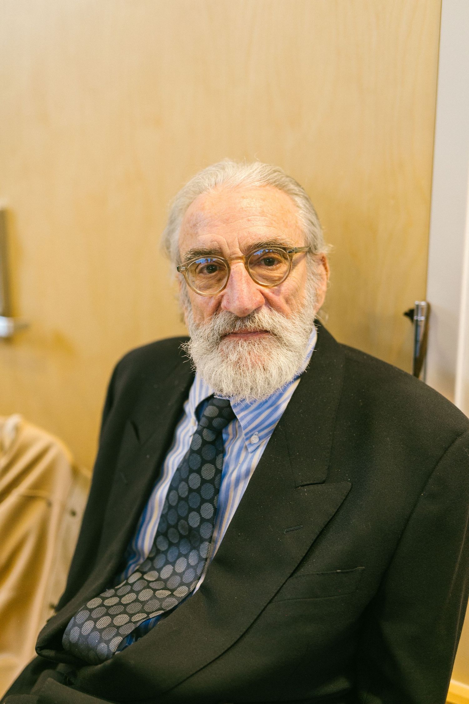 Senior gentleman in business attire wearing glasses and a polka dot tie seated against a wooden wall.