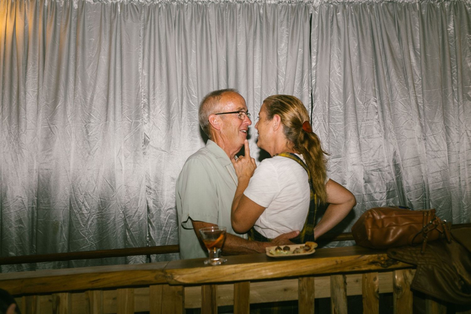 Tender romantic moment shared between two people sitting on a wooden bench against a silver curtain backdrop.