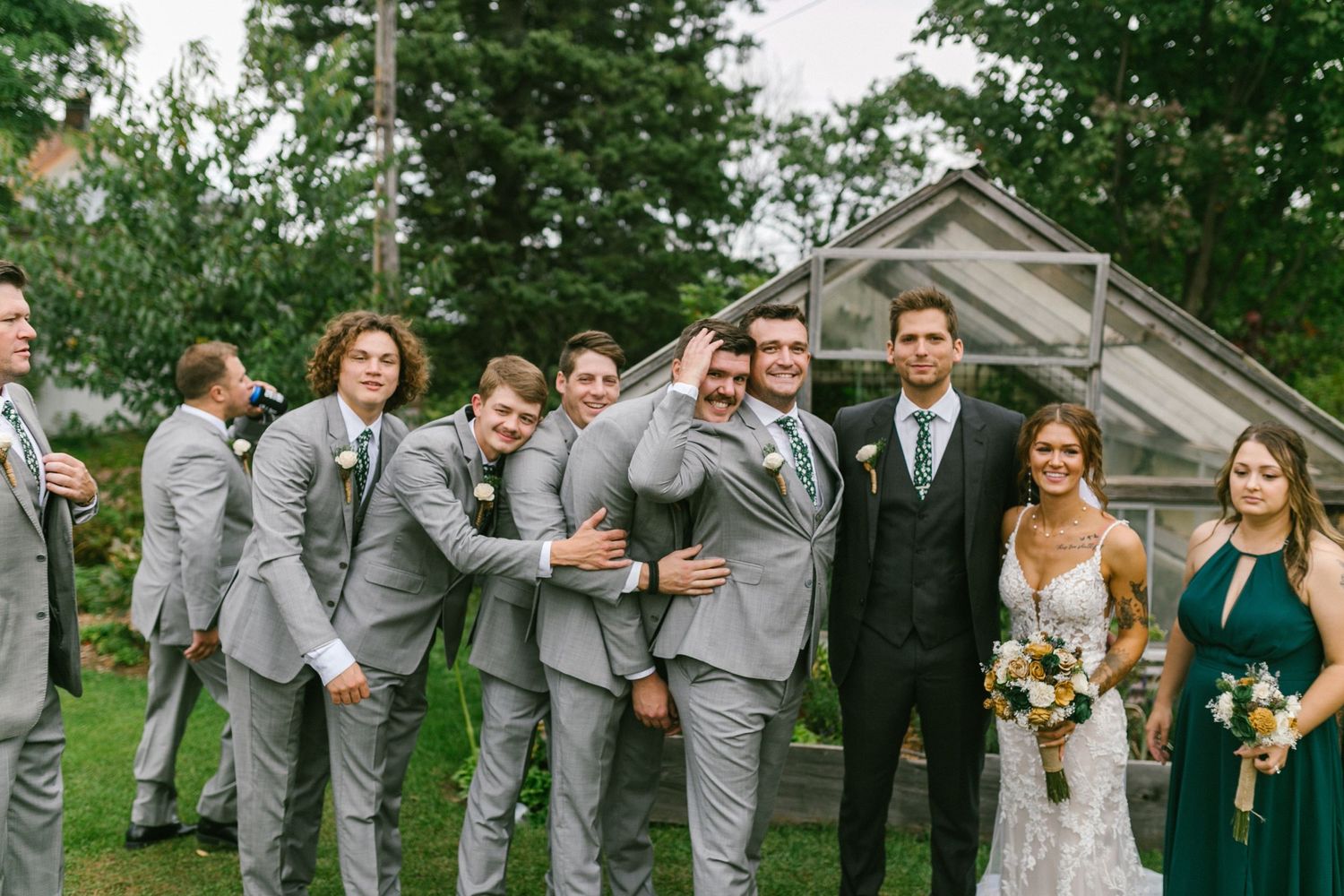 Wedding party in coordinated light grey suits and formal attire pose playfully on a green lawn.