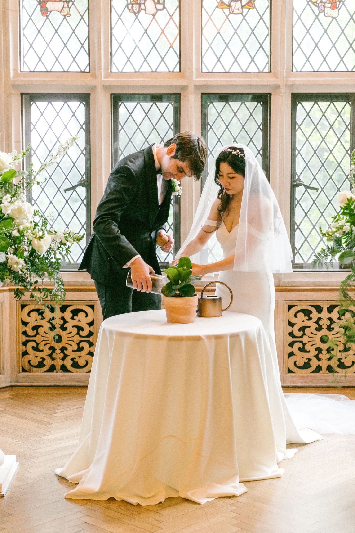 Bride and groom perform a unity plant watering ceremony before ornate leaded glass windows with white florals.