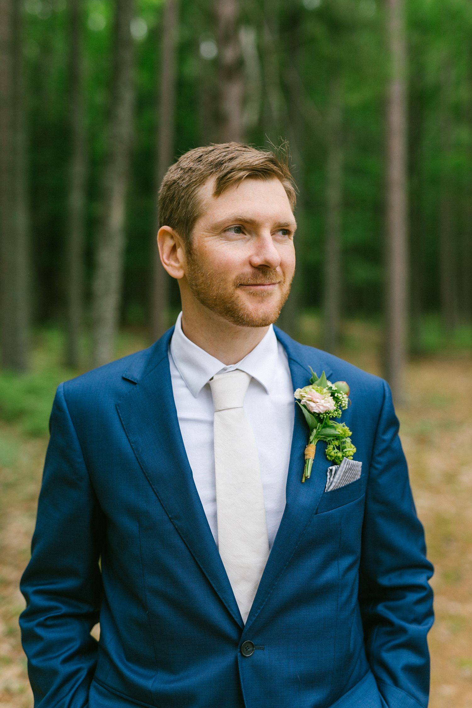 A groom wearing a navy blue suit with a white tie and floral boutonniere stands in front of a wooded forest background.