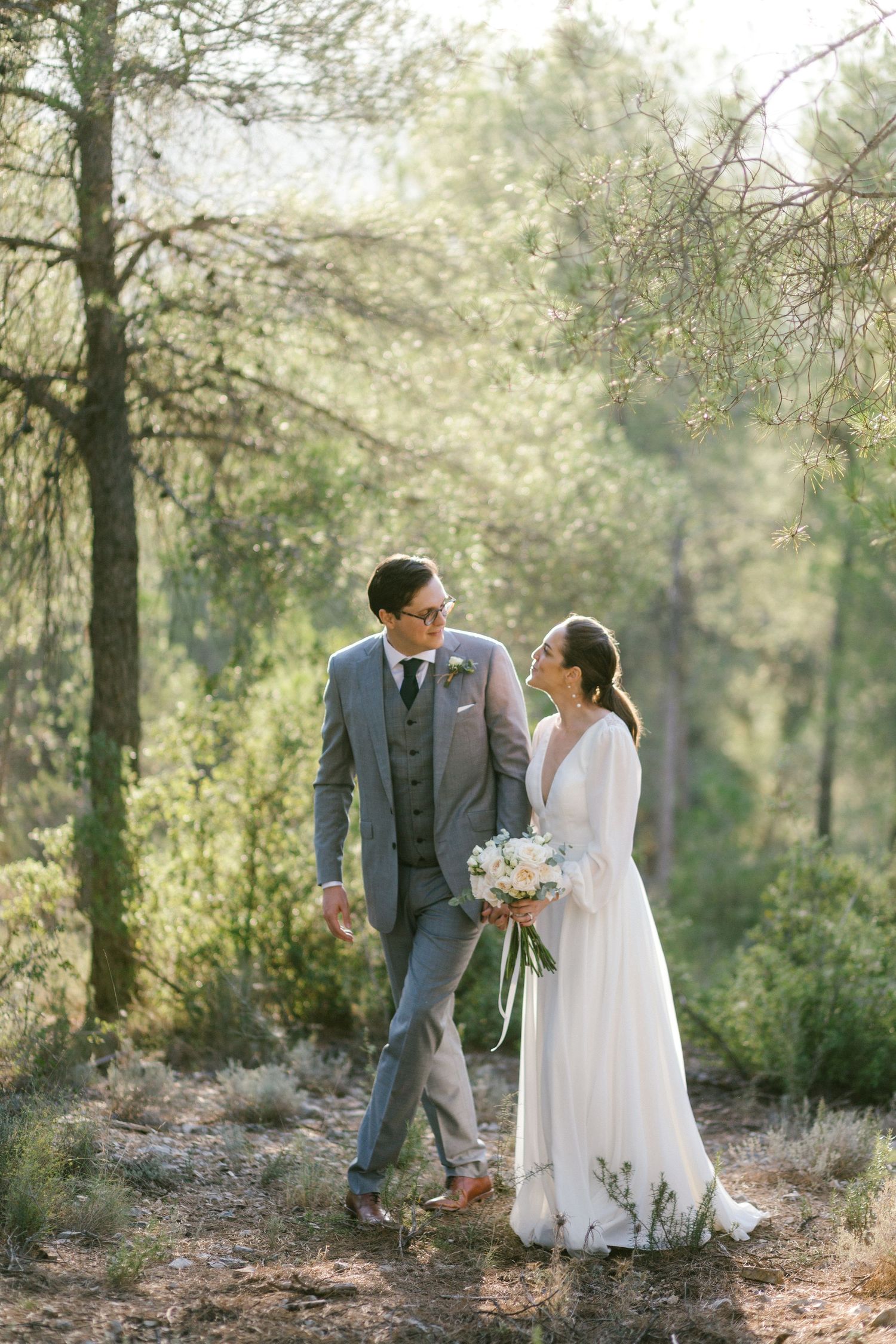 A romantic wedding couple stands together in a sunlit forest setting, with the bride holding a delicate bouquet of flowers in Spain.