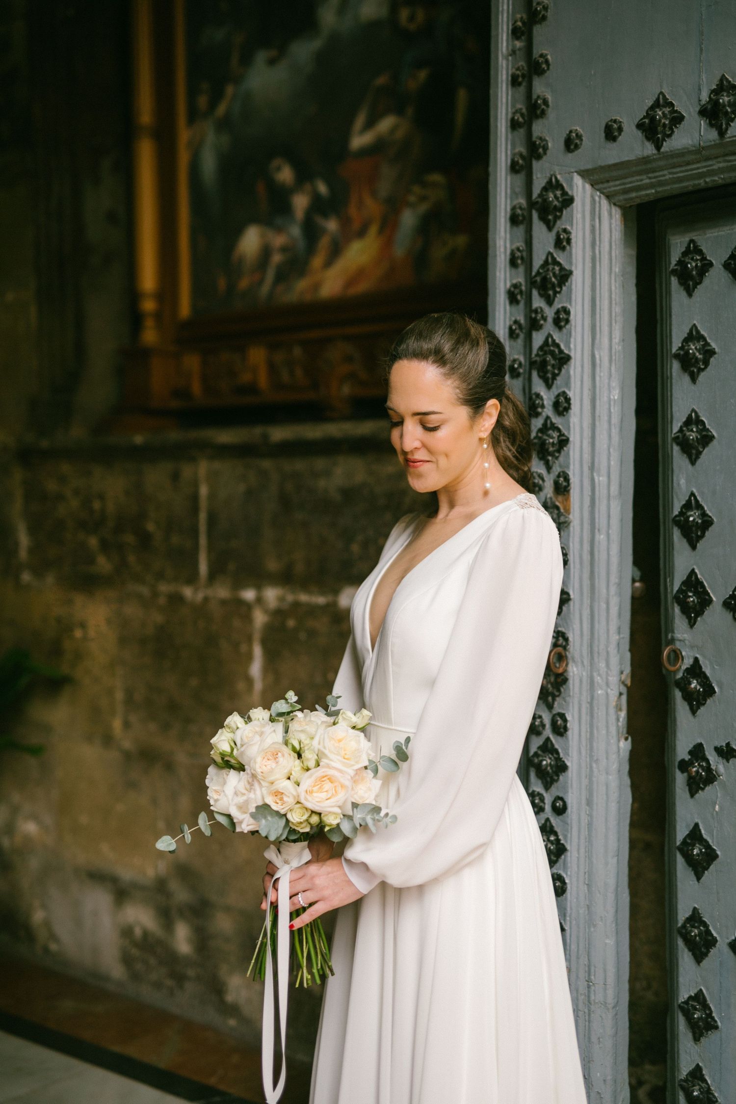 An elegant bride in a long-sleeved white gown with billowing sleeves holds a soft pastel bouquet by an ornate doorway.