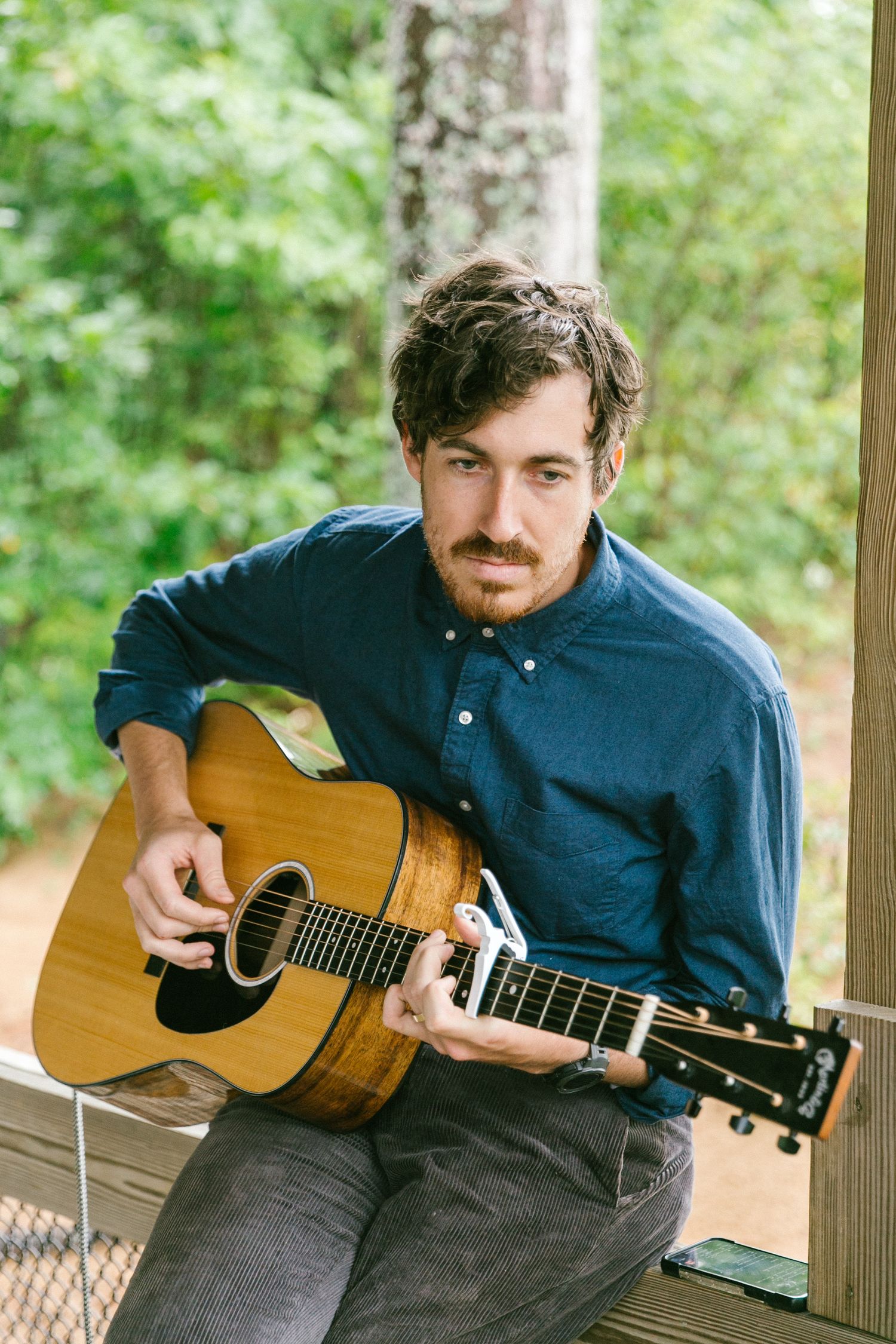 Musician playing acoustic guitar while seated on outdoor porch.