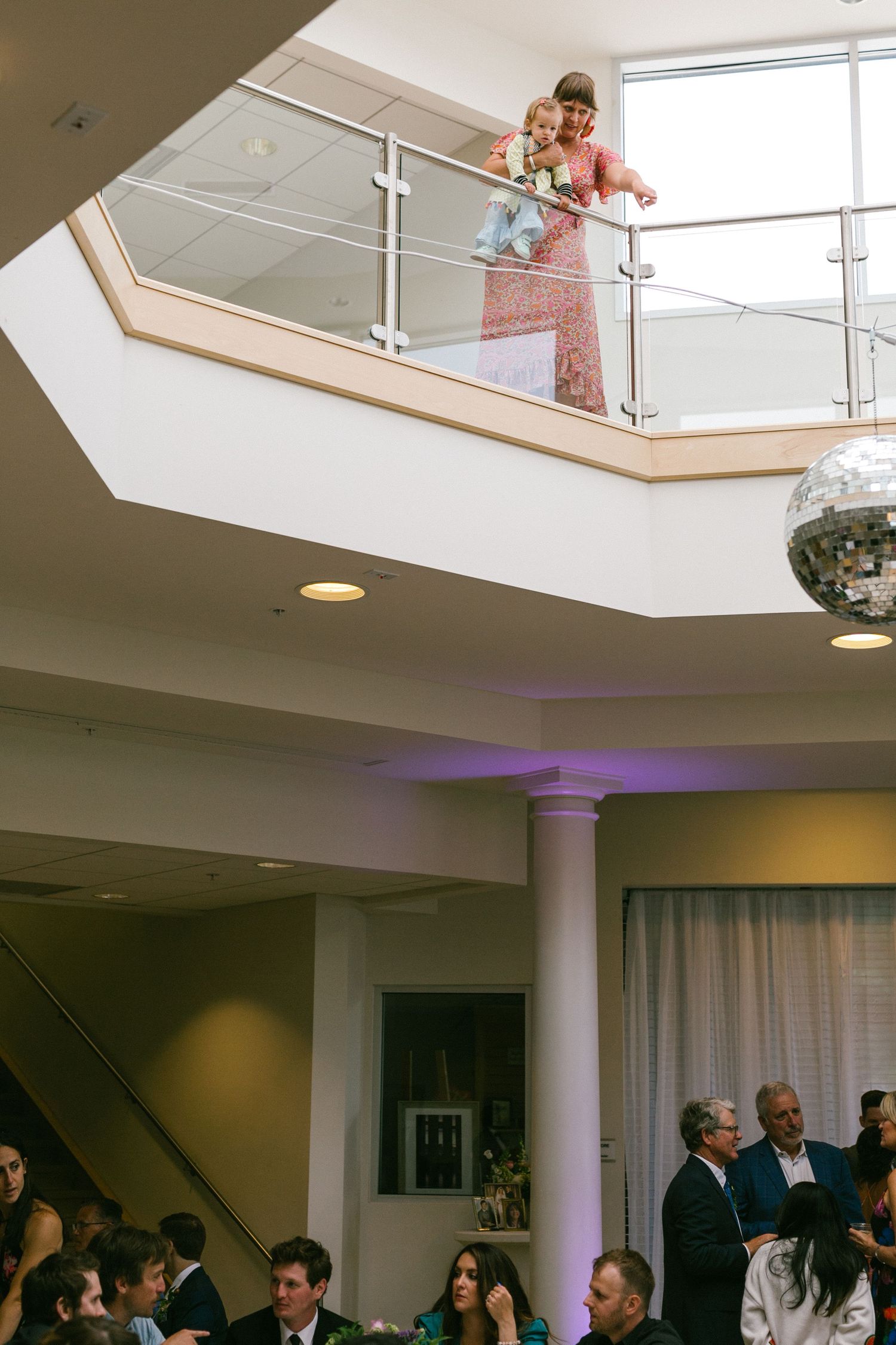 A person in a pink dress looks down from an interior balcony with a disco ball hanging below at an event space.