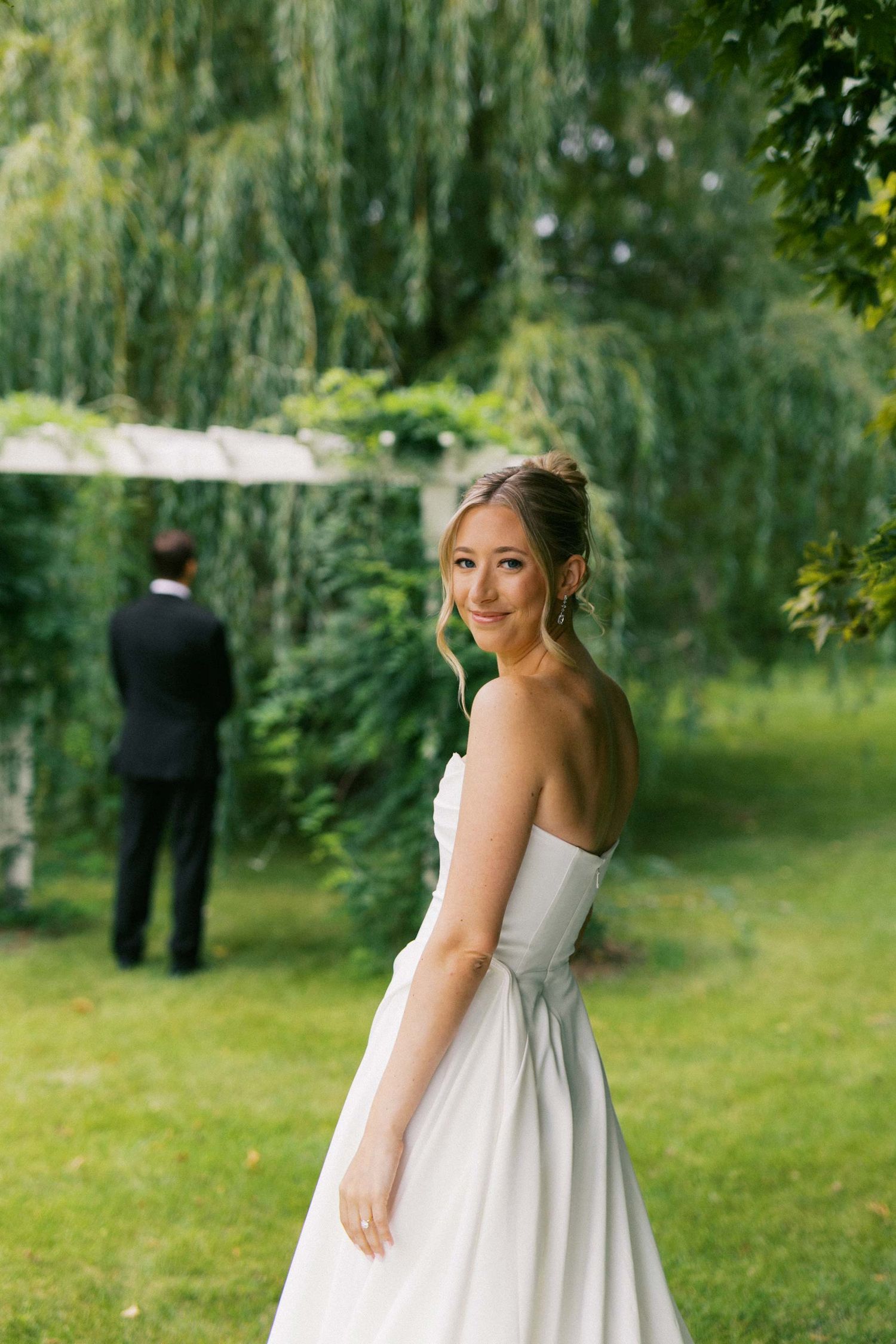 A bride in a white strapless wedding dress poses in a garden with willow trees while her groom stands in the background.