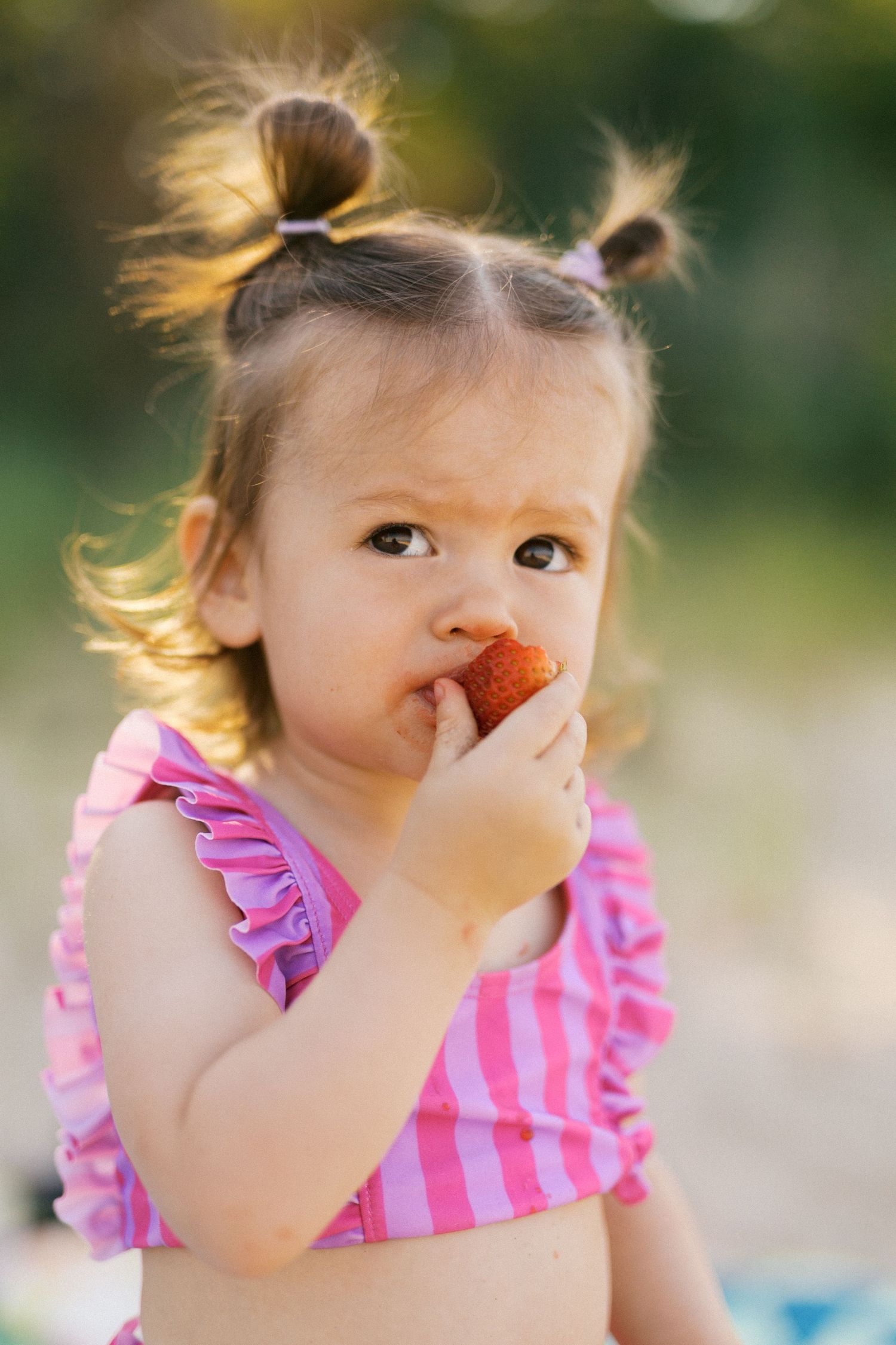 Cute toddler girl with pigtails in pink swimsuit eating a fresh strawberry outdoors in summer.