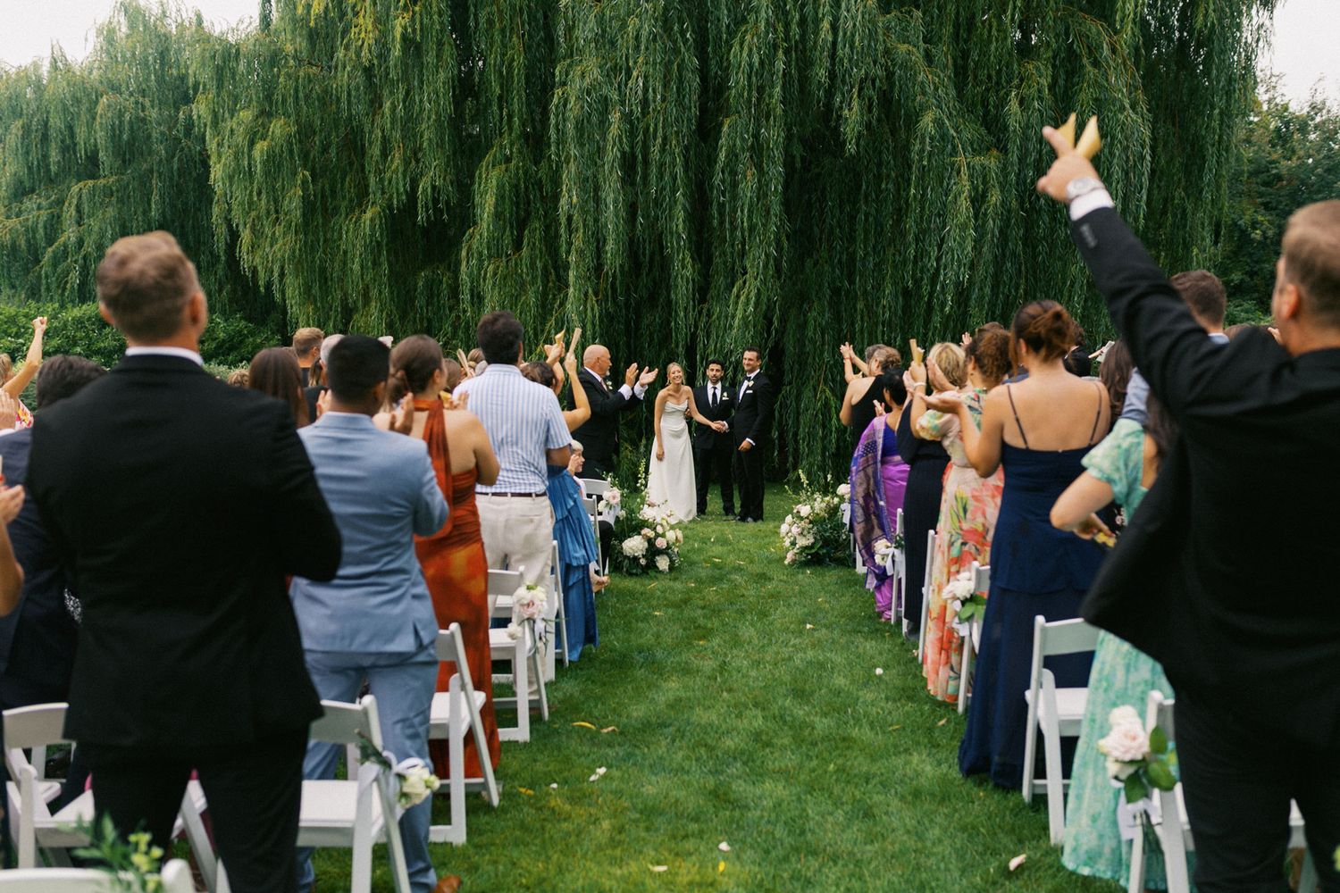Wedding guests cheer as bride and groom walk down a lush outdoor aisle beneath a weeping willow tree.