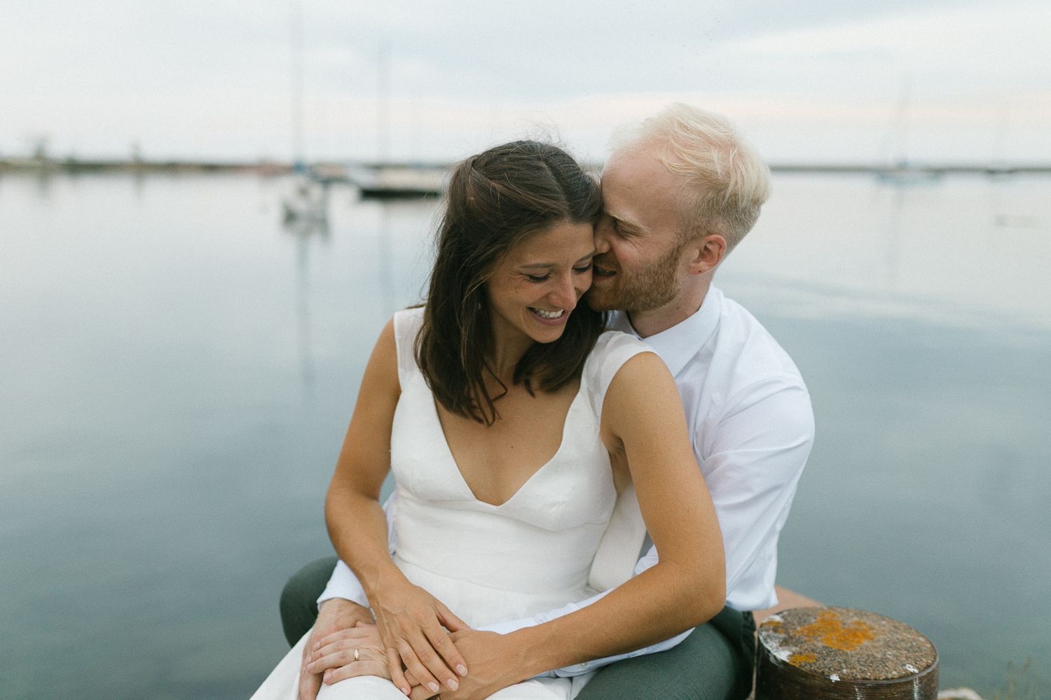 Romantic couple sitting together by calm waterfront with sailboats, man nuzzling woman in white dress.