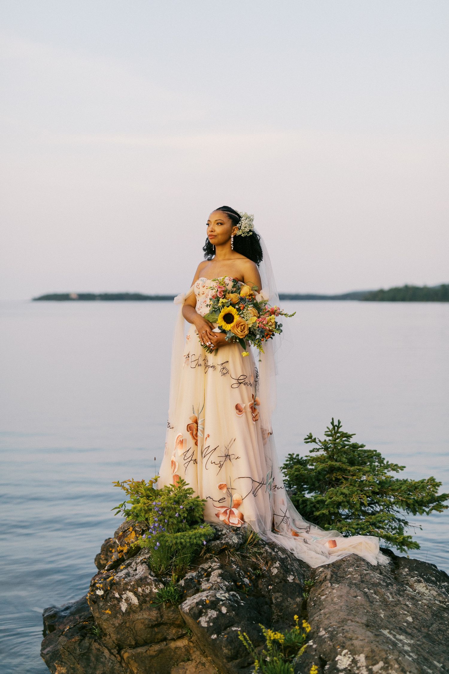 Elegant bride in a floral gown holding a sunflower bouquet while standing on a rocky lakeside cliff.