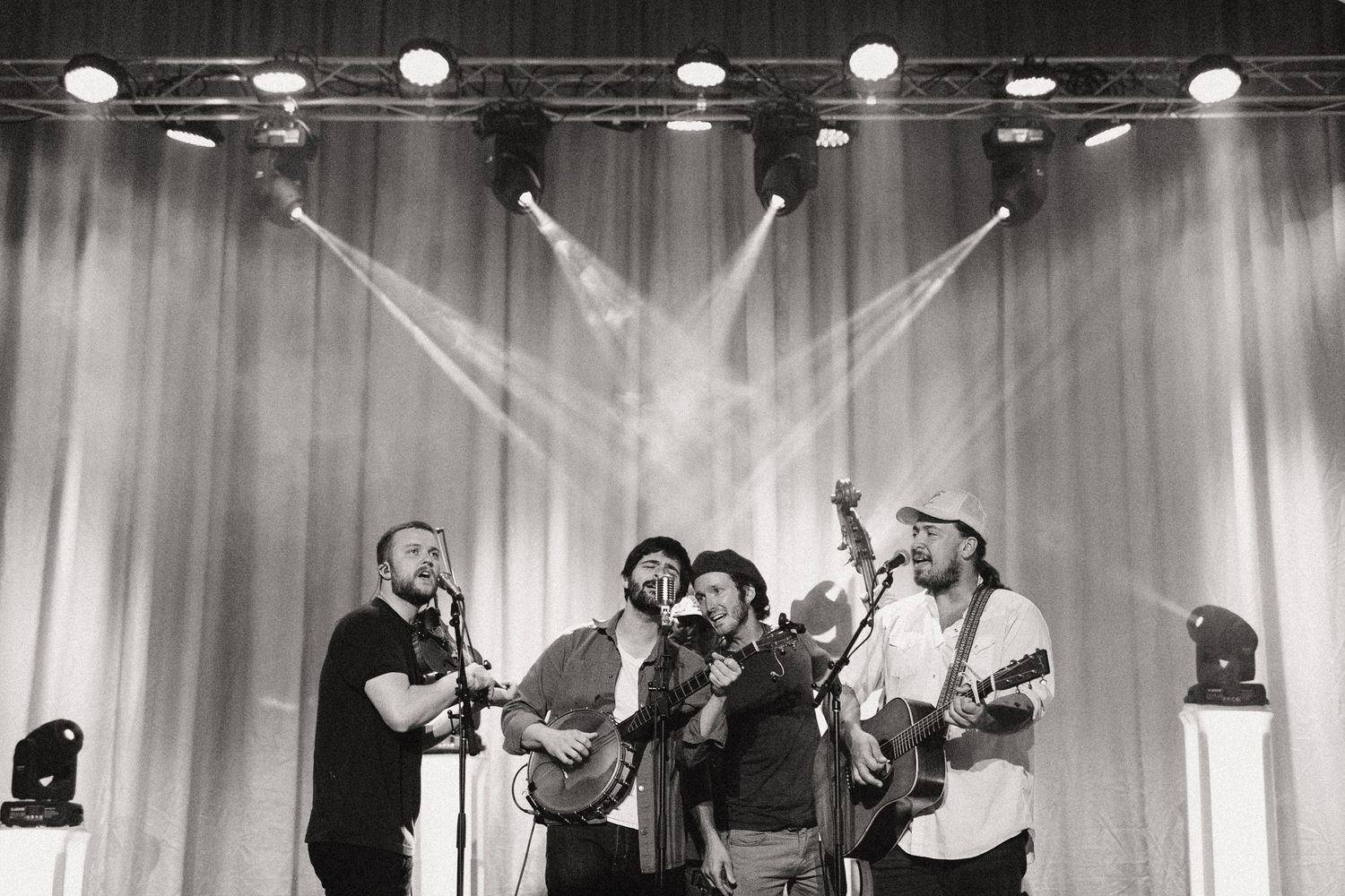 A black and white photo shows musicians performing on stage with dramatic backlighting and spotlights creating beams through haze.