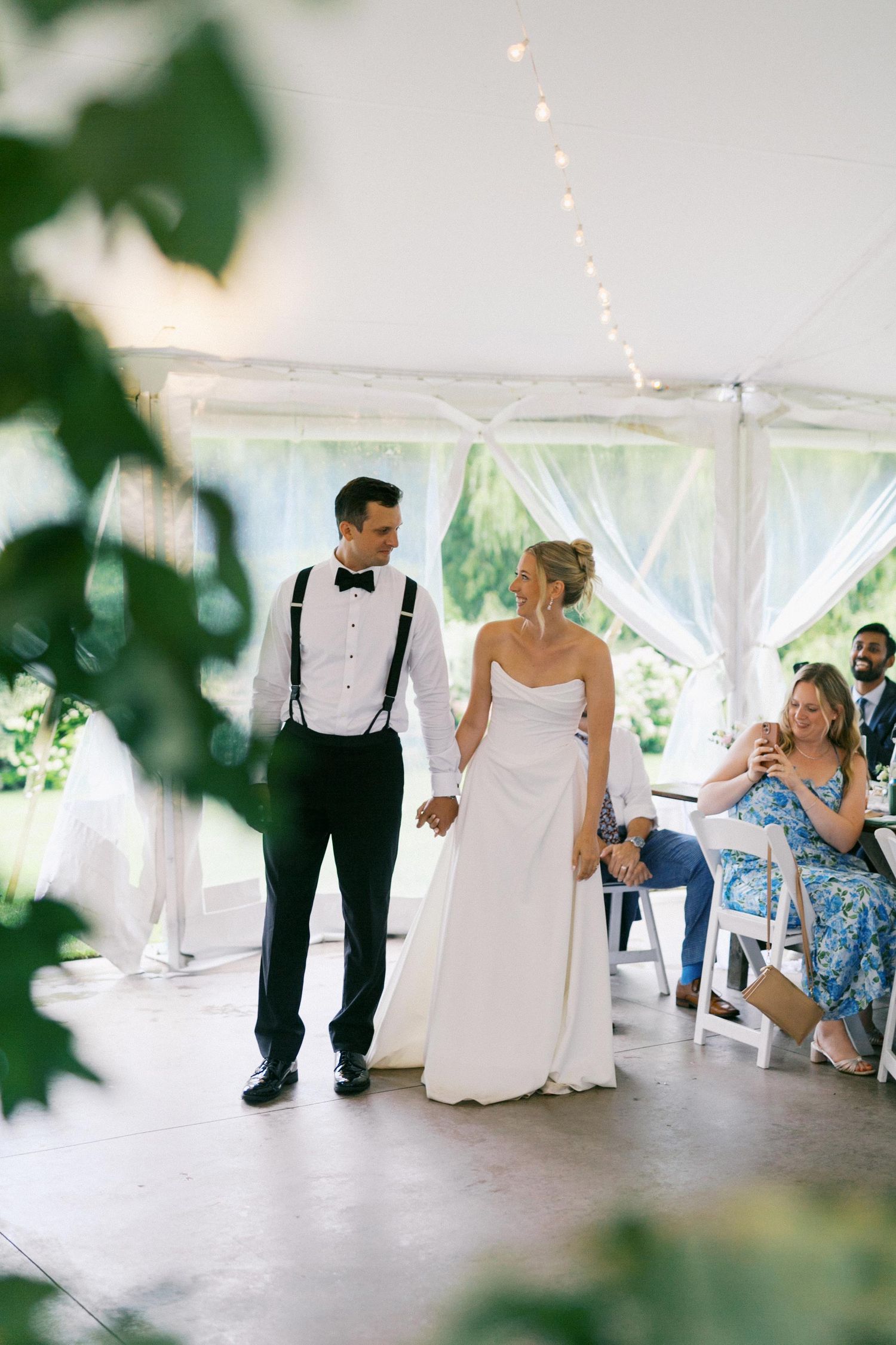 A couple walks together under a white tent decorated with string lights at an outdoor wedding reception.