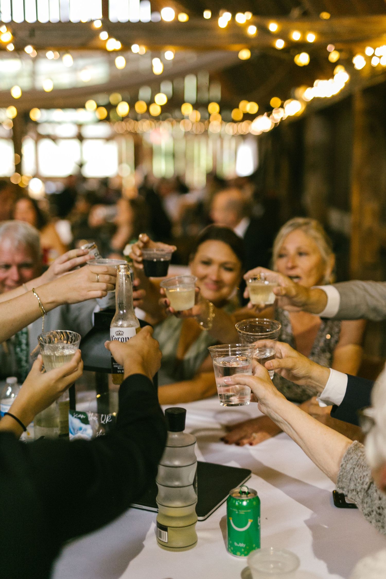 Group toasting with drinks at a dimly lit wedding reception with string lights overhead.