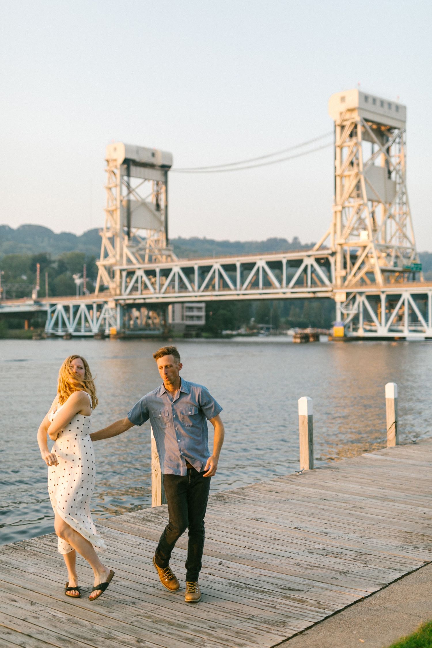 Couple dancing on wooden dock with historic lift bridge spanning across the river behind them.