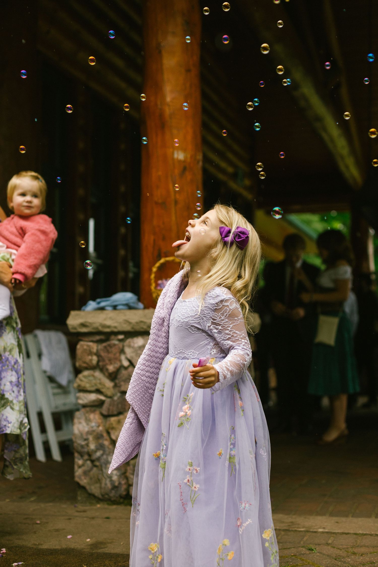 A young person in a purple dress chases bubbles at an outdoor venue while someone watches in the background.