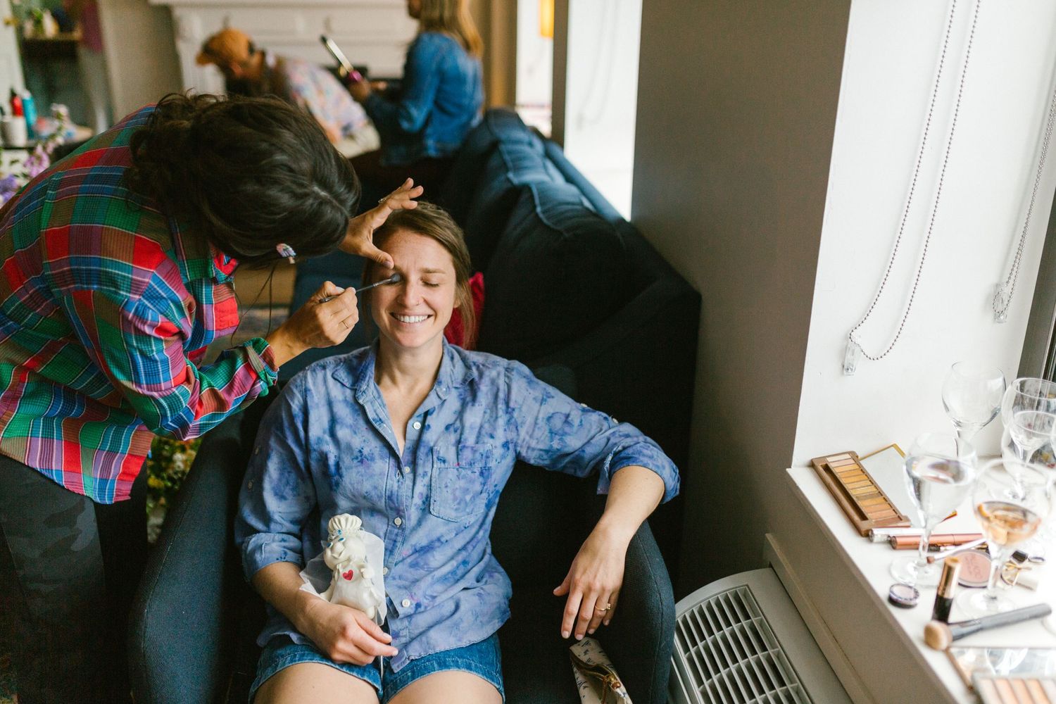Two people sitting indoors getting ready with makeup supplies and hair styling tools.
