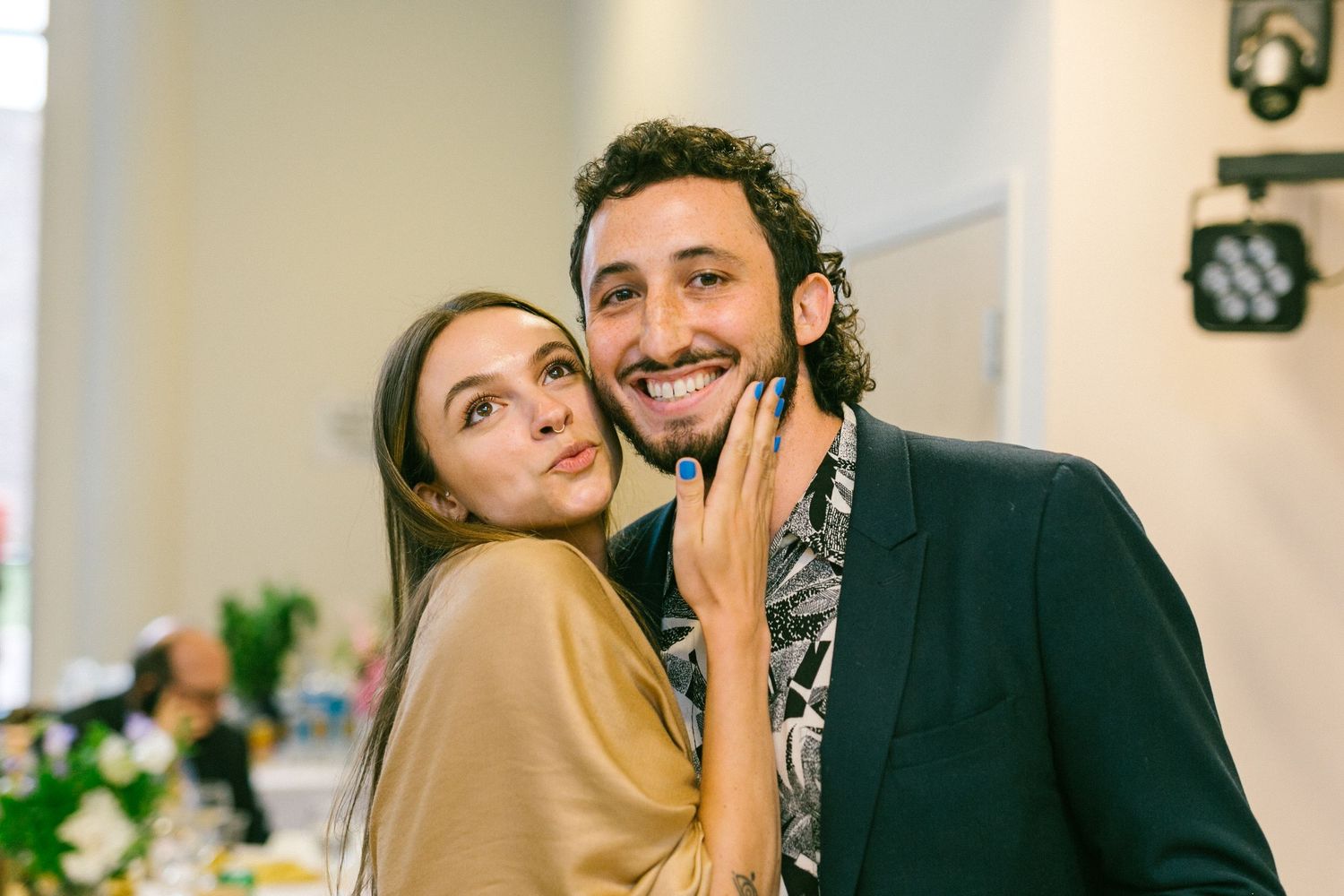 A couple embraces and smiles warmly while standing in an indoor setting with a clock on the wall.