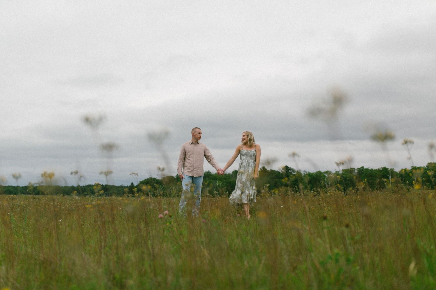 Two people walk together through a misty green field beneath cloudy skies.