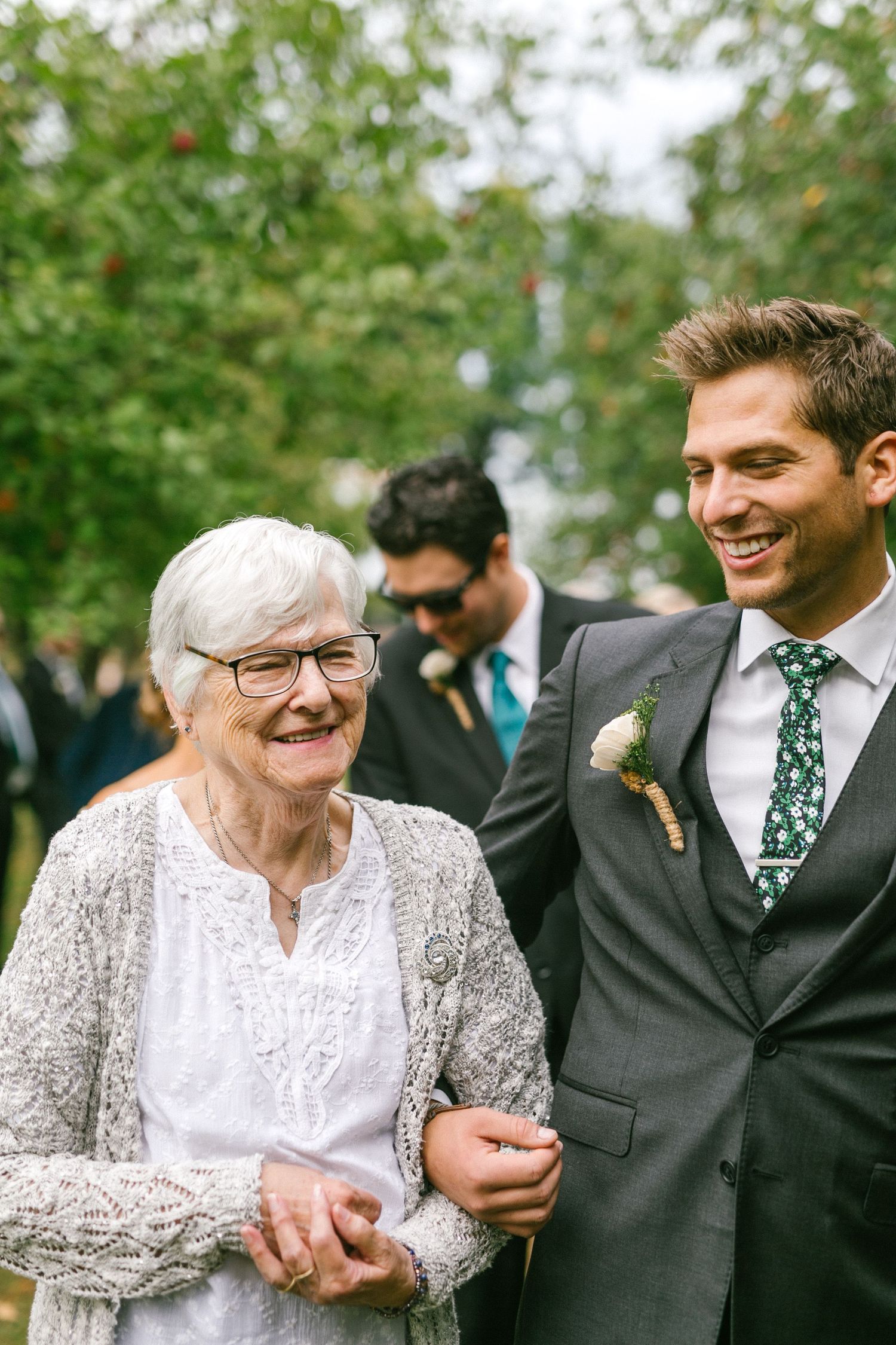 Happy elderly Grandma in white lace dress arm-in-arm with groom in grey suit at outdoor wedding celebration.