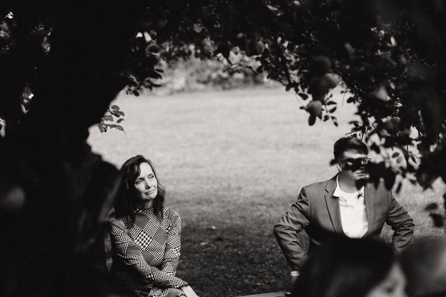 A black and white photo shows two people sitting together framed by overhanging tree branches.