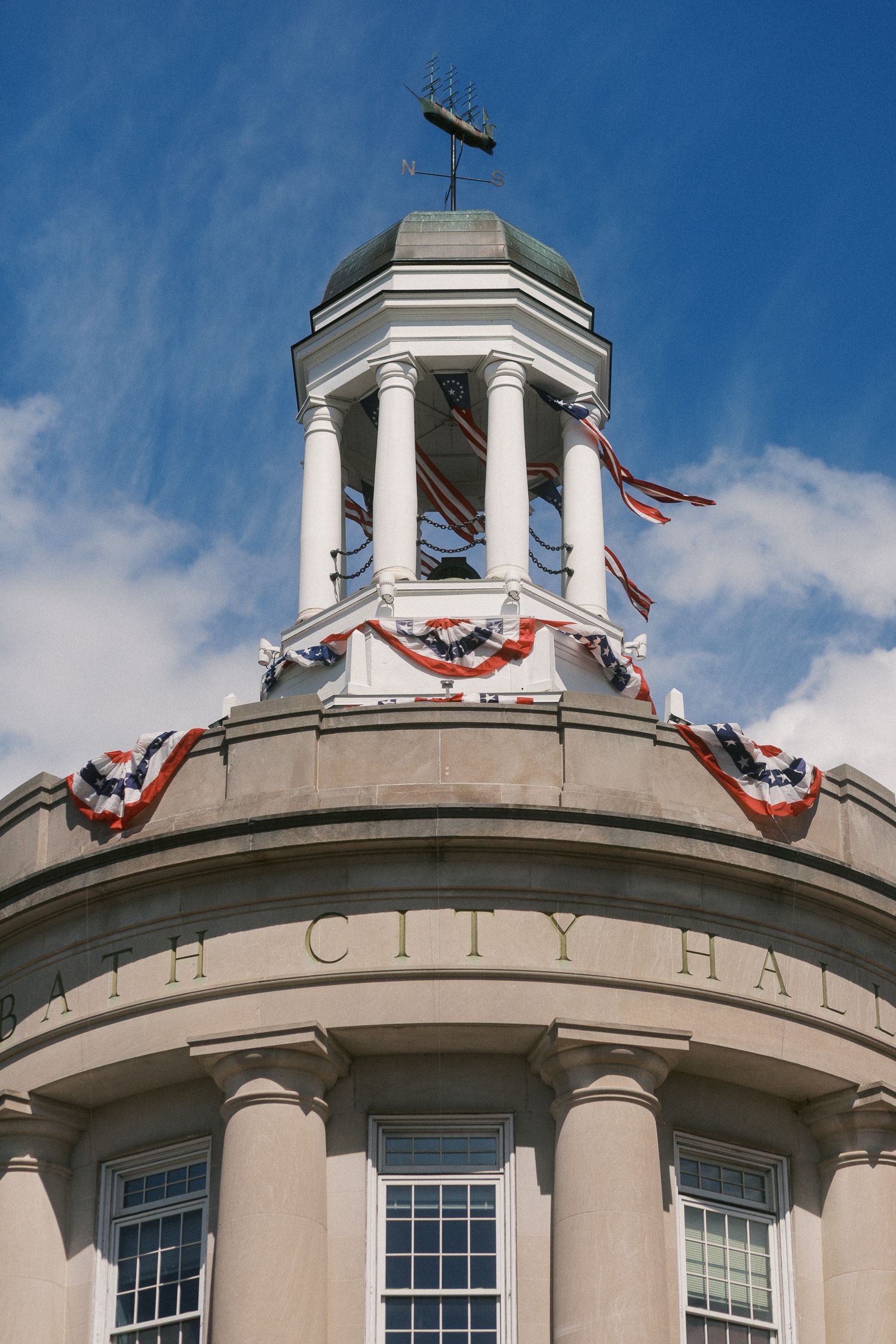 Historic city hall building with white cupola, patriotic bunting decorations, and weather vane against a blue sky.