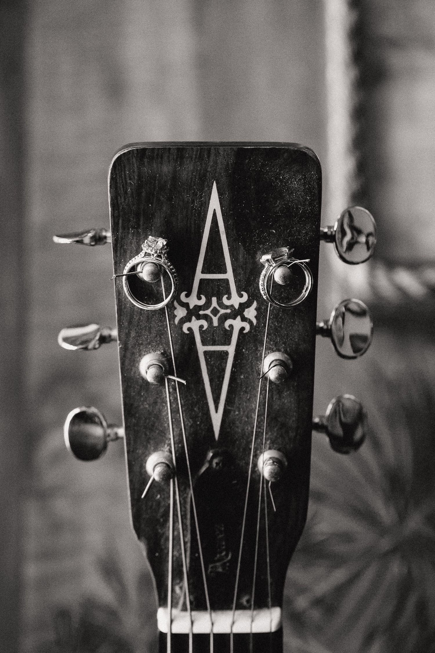 Black and white photo of acoustic guitar headstock with two wedding rings resting on the tuning pegs.