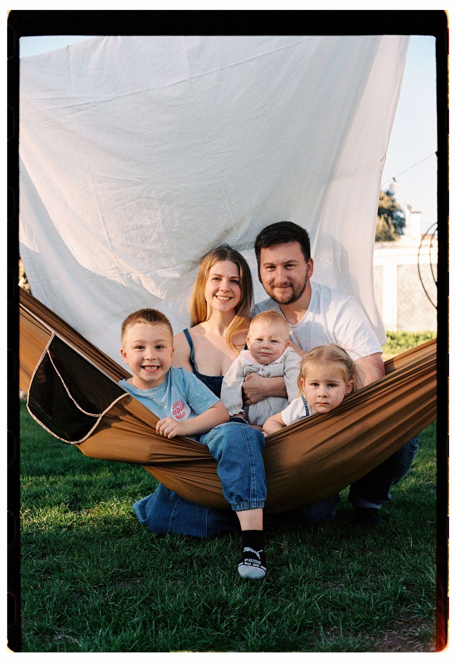 Young family of five smiling together in a brown hammock outdoors on green grass with white fabric backdrop.