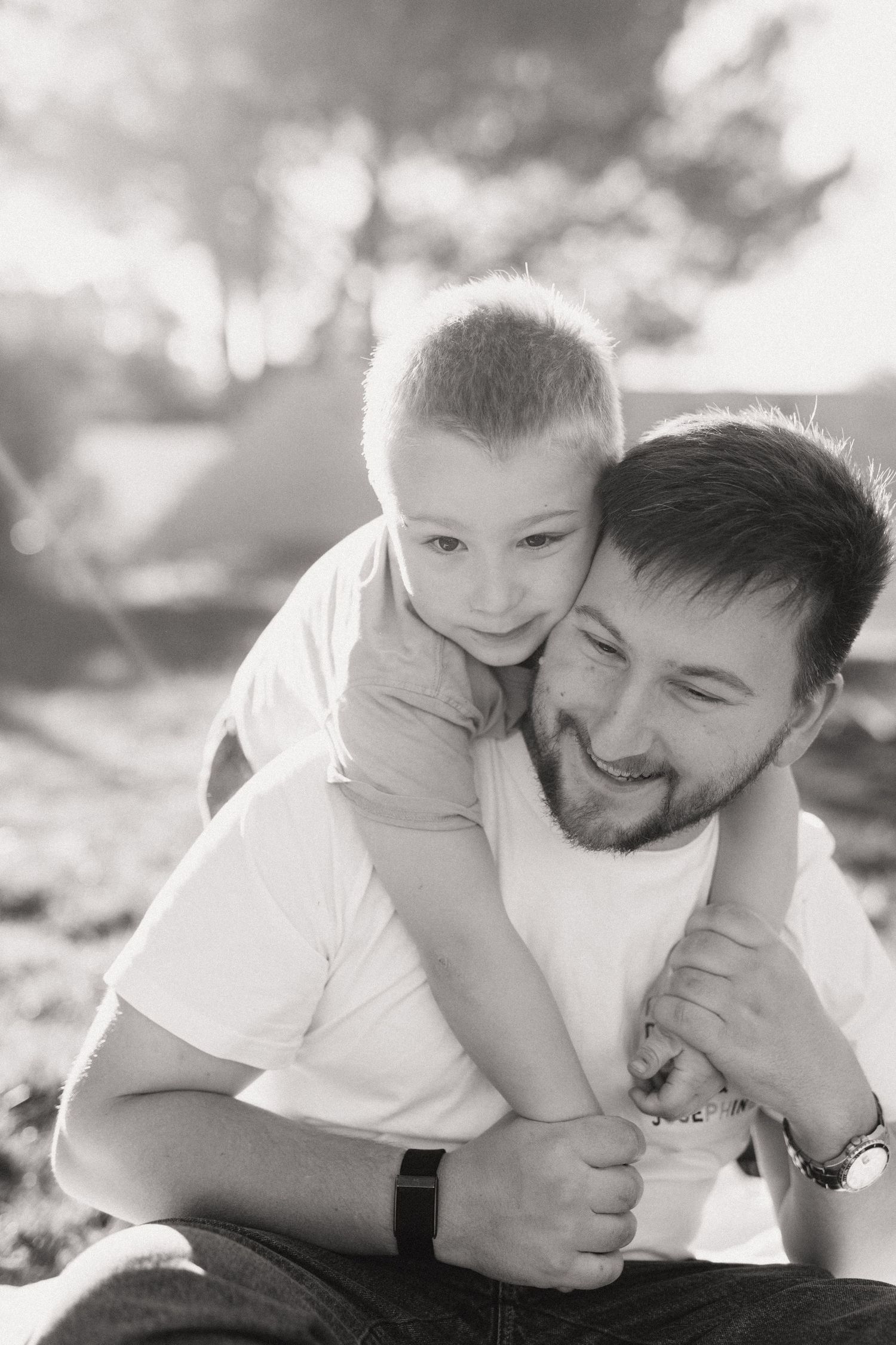 Black and white photo of a smiling father and young son sharing a warm embrace outdoors.