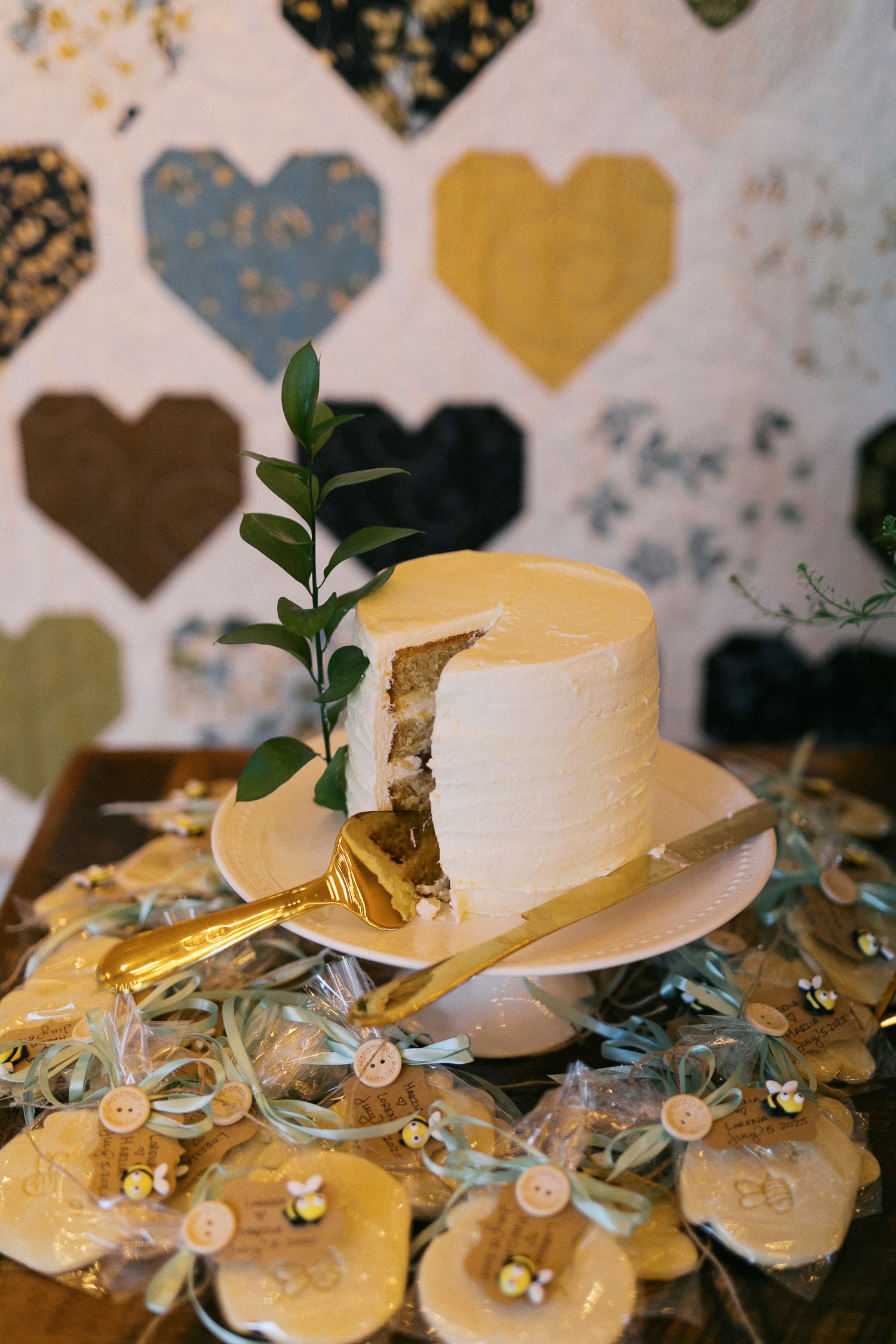 White frosted wedding cake with a slice cut out, gold server, greenery sprig, honey favors, and heart quilt backdrop.