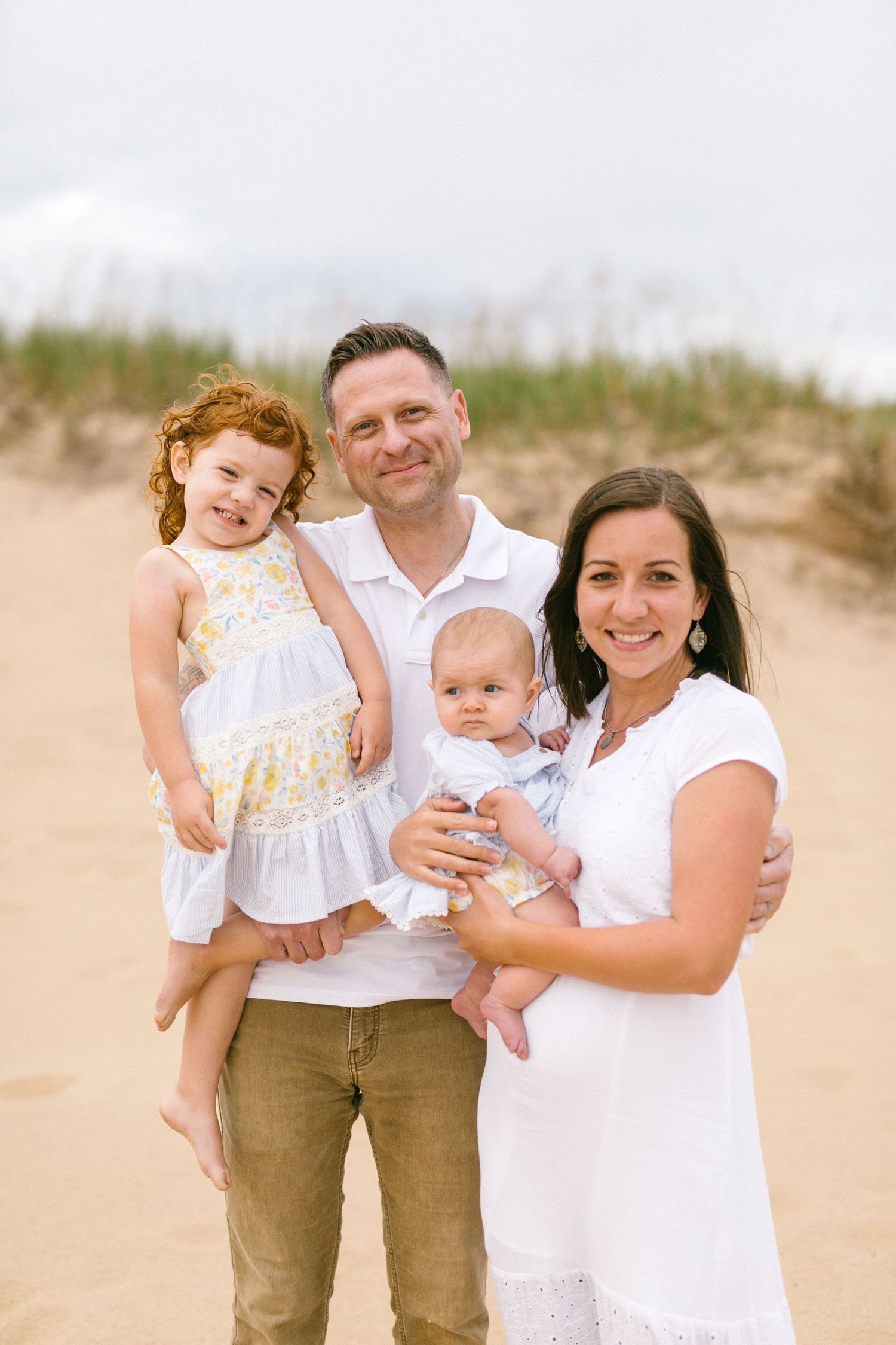 Happy family of four posing on a sandy beach, parents holding two young children in matching outfits.
