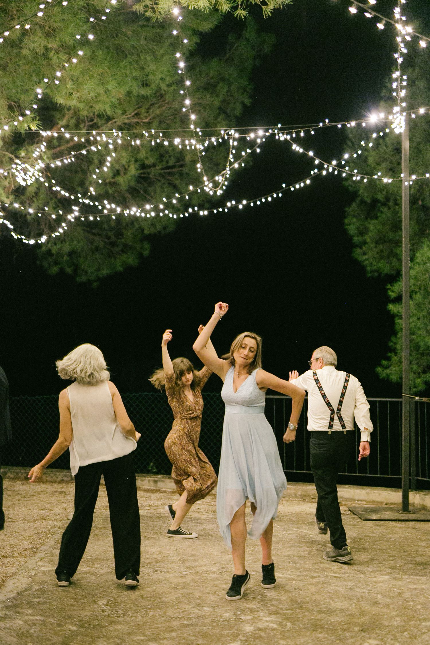 Guests dance outdoors under string lights at a nighttime wedding reception surrounded by trees.
