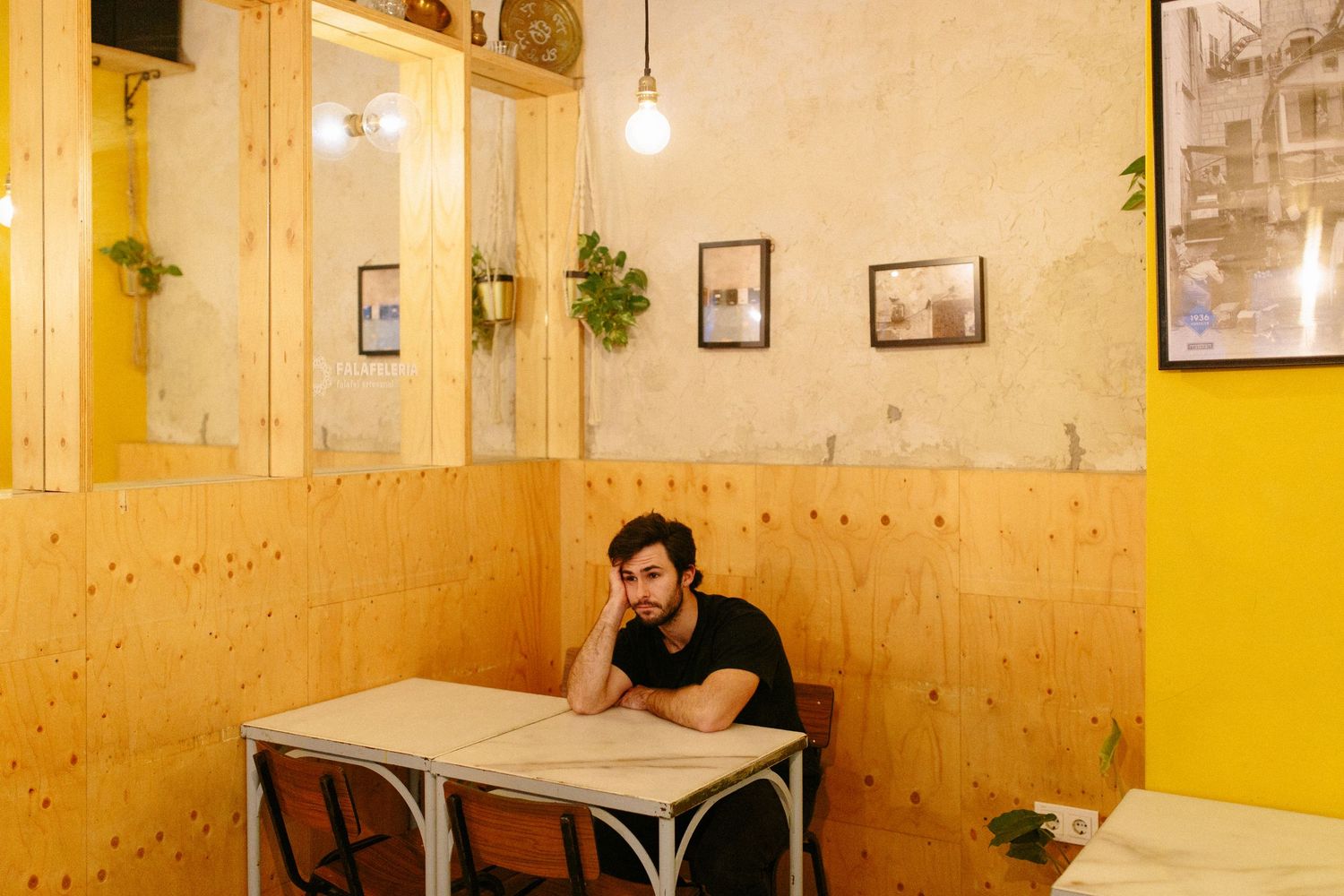 Young bearded man sitting alone at a table in a yellow-walled cafe.