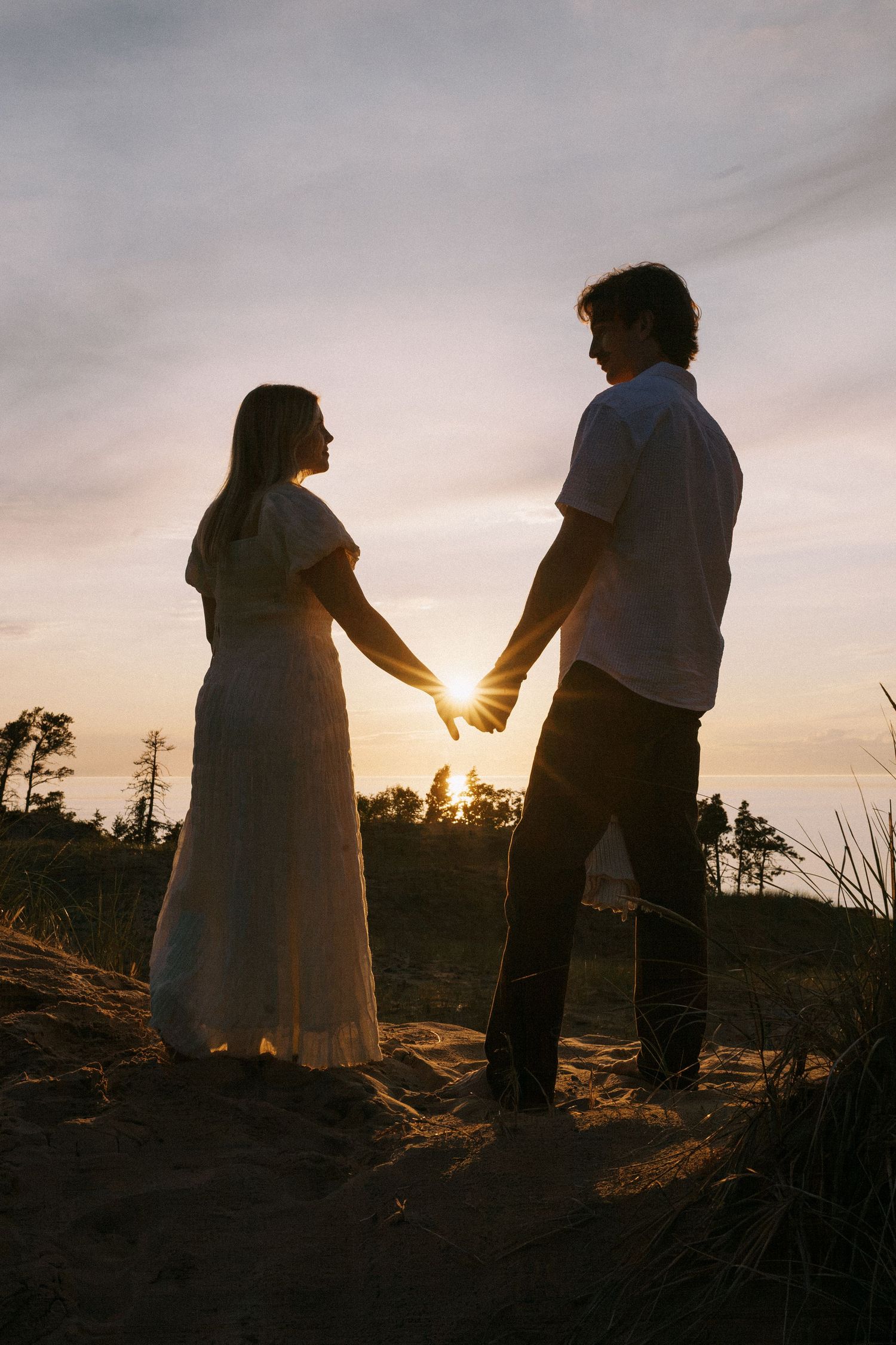 Couple holding hands at sunset on sandy dunes, silhouetted against golden sky, woman in white dress.