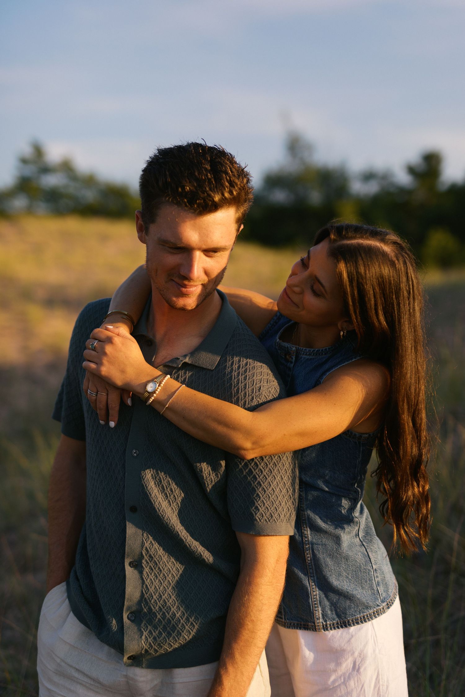 Young couple embracing in a golden field at sunset, woman hugging man from behind.
