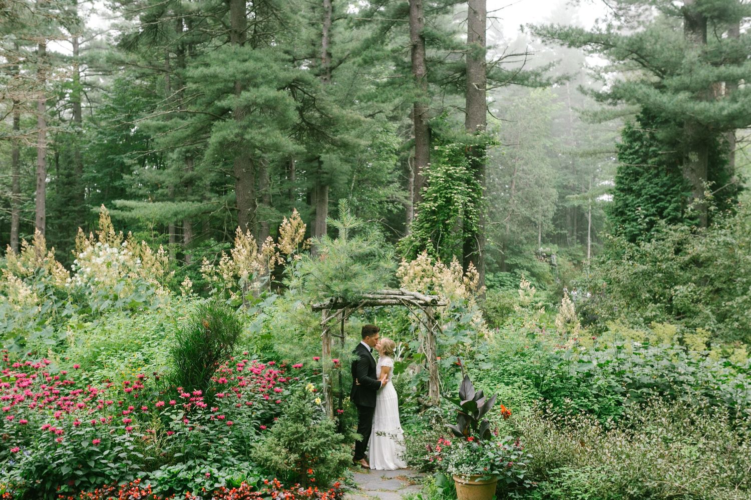 A bride and groom kiss on a lush garden with blooming flowers and tall evergreen trees around them at their Marquette, MI wedding.