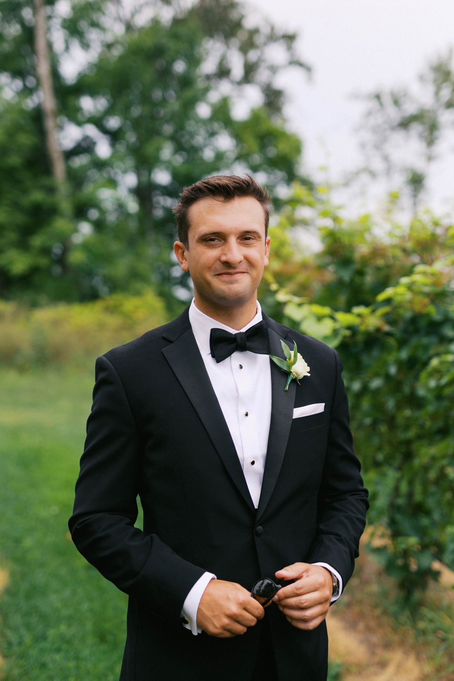 Groom in classic black tuxedo and bow tie adjusts his jacket while standing in a lush outdoor garden setting.