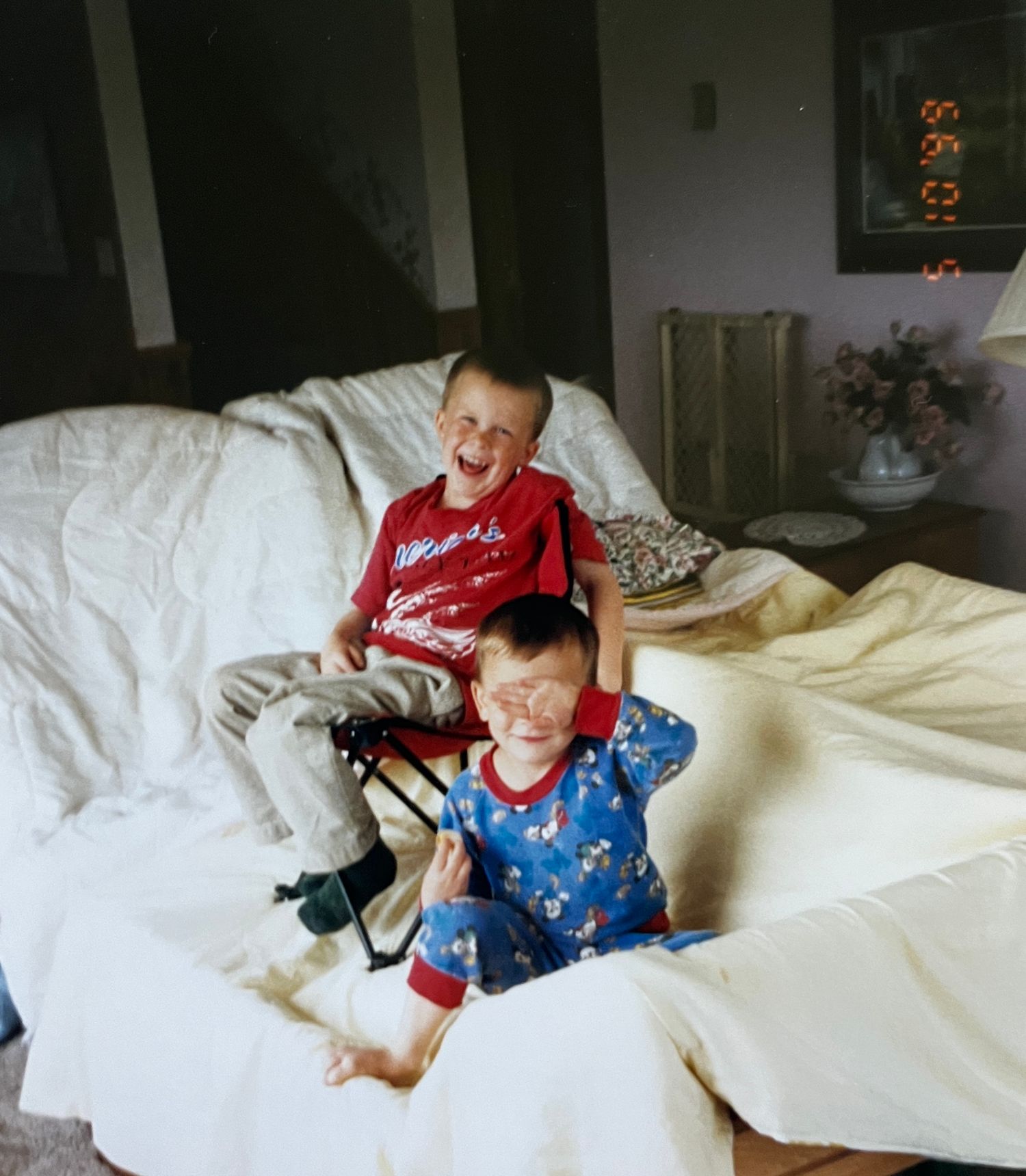 Two young boys laughing and playing on white couch in living room, wearing colorful pajamas.