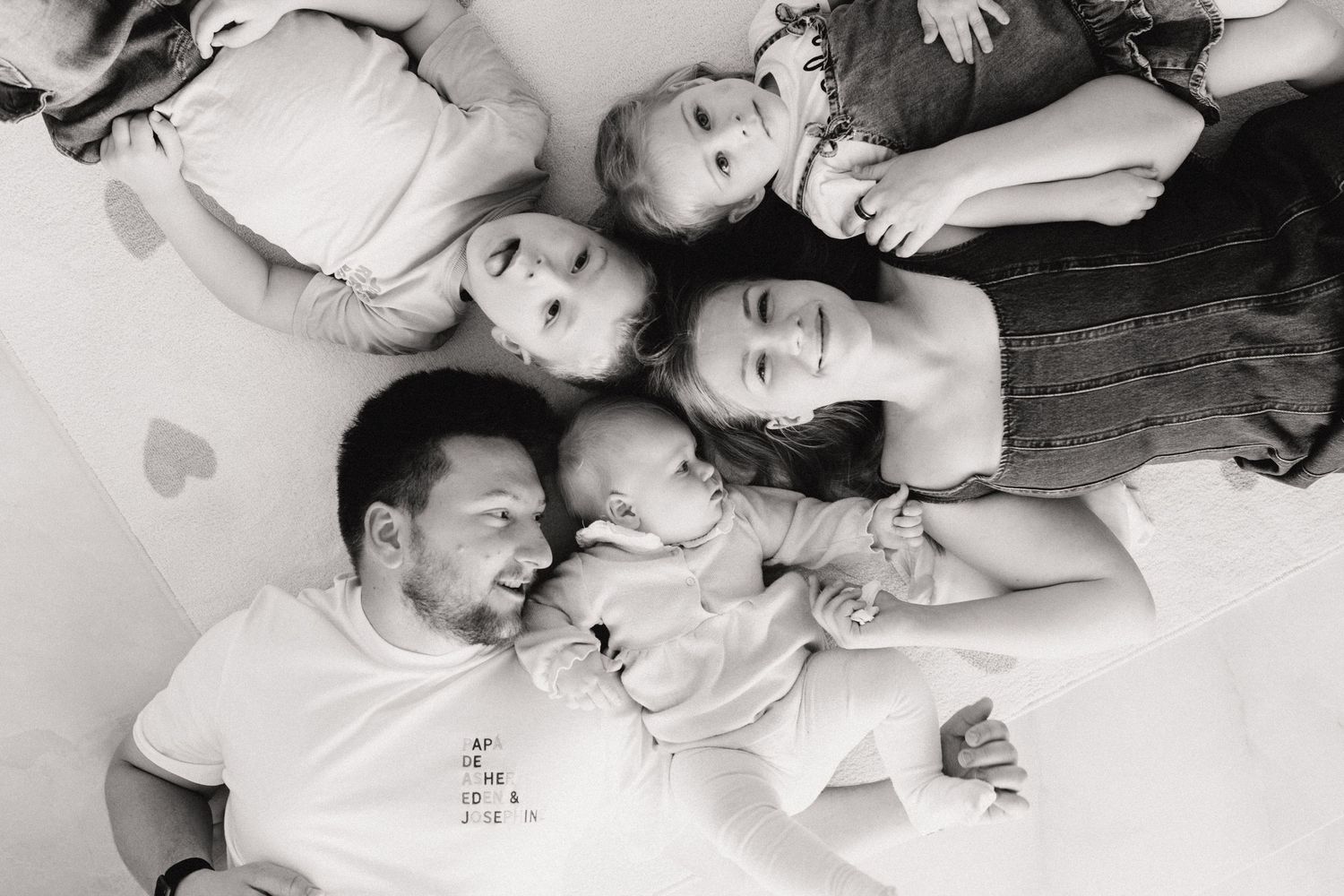 Black and white overhead photo of family of five lying together on a white surface.