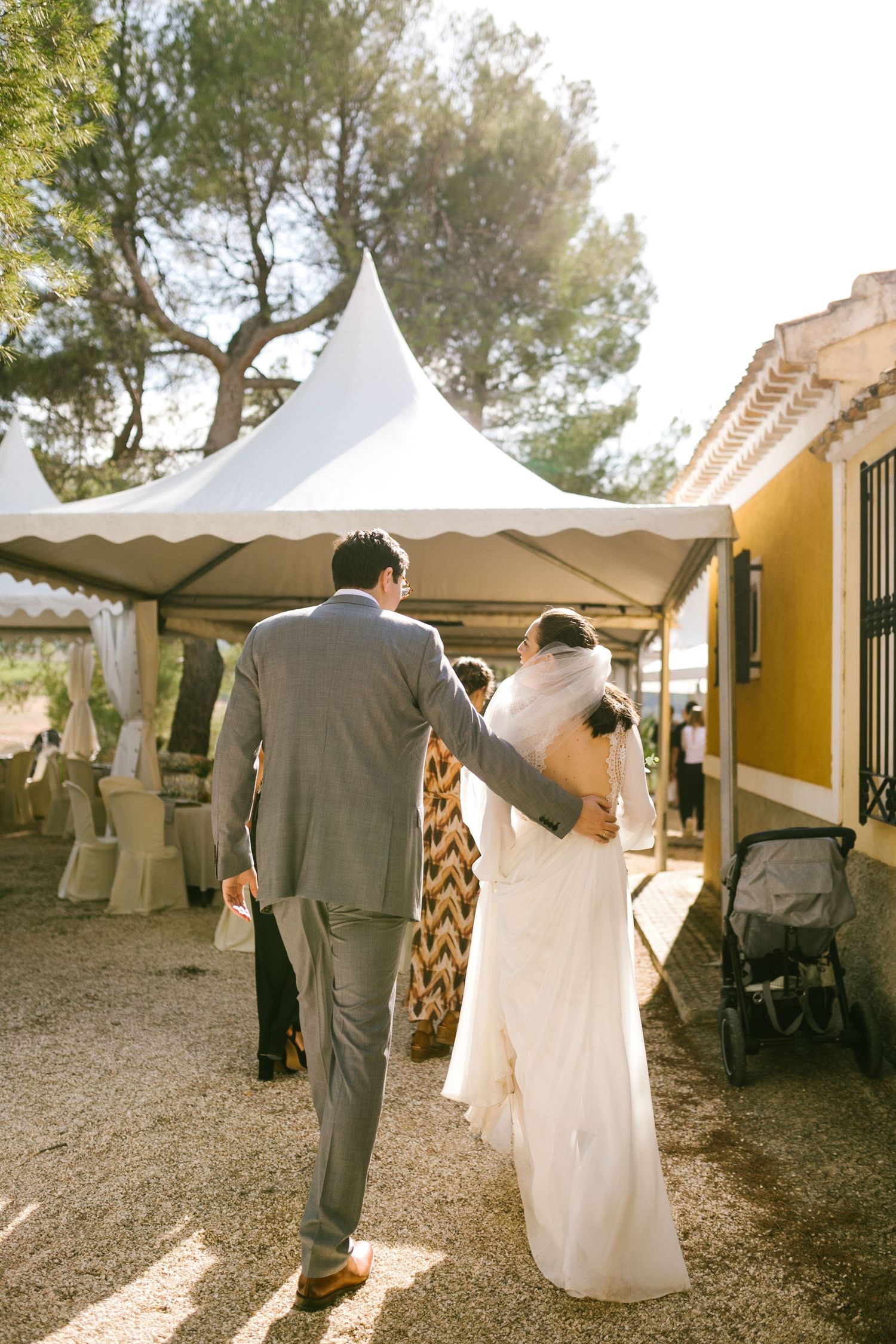 A couple walks under an elegant white tent canopy at an outdoor wedding reception on a sunny day.