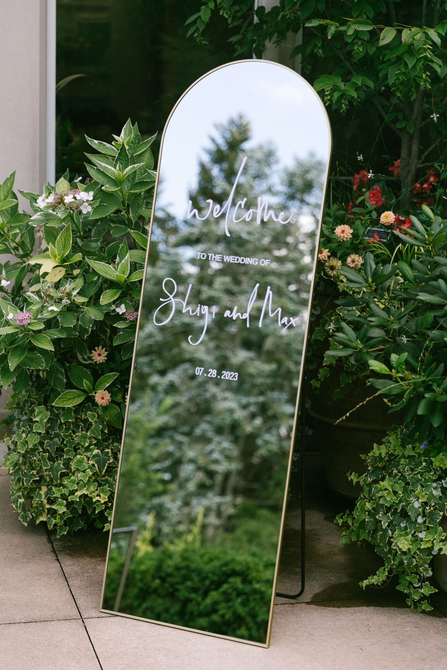 Elegant arched mirror wedding welcome sign for Shaya and Max, dated 07.28.2023, surrounded by lush greenery.