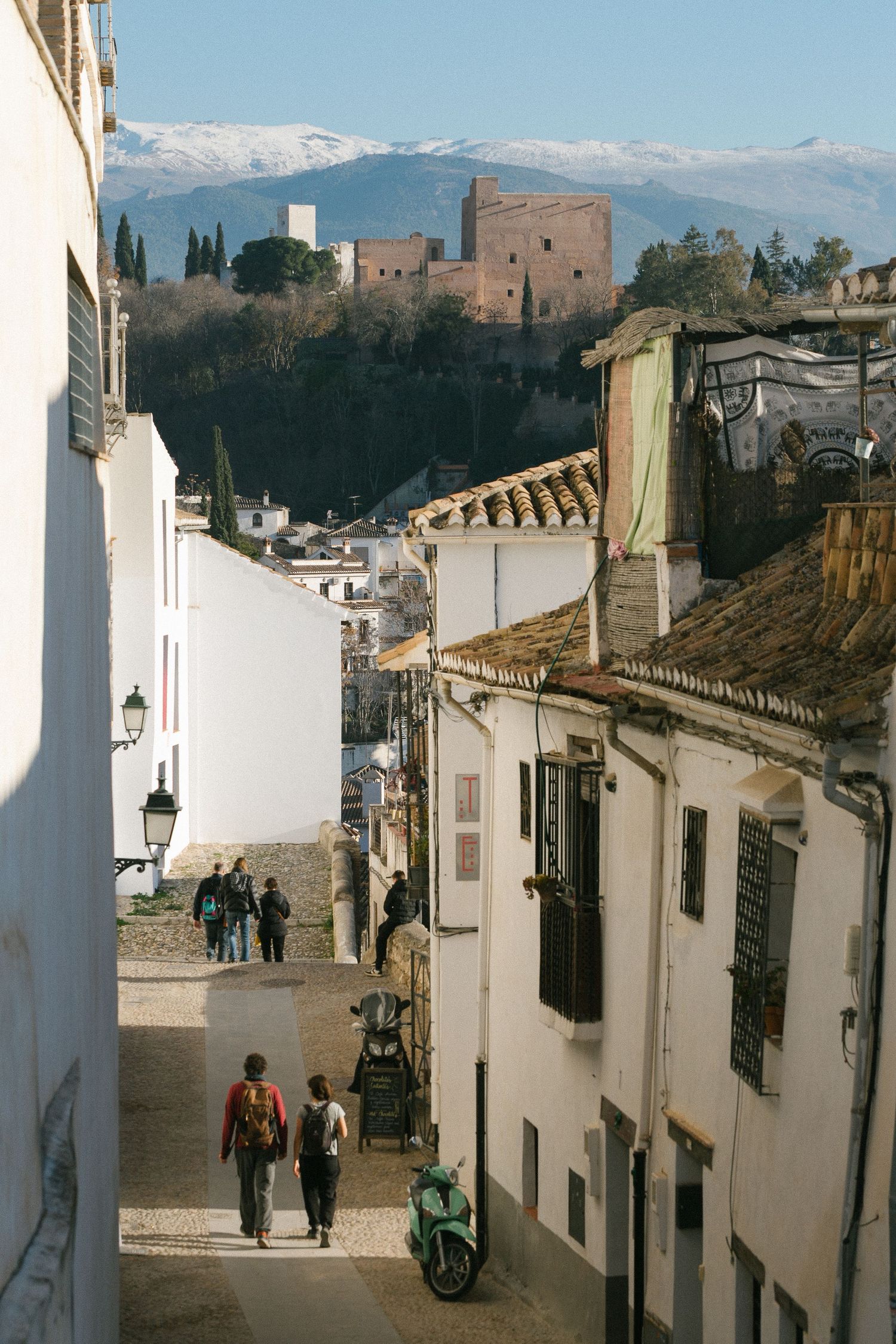 A steep cobblestone street in Granada, Spain with white buildings and the Alhambra fortress visible on the hilltop.