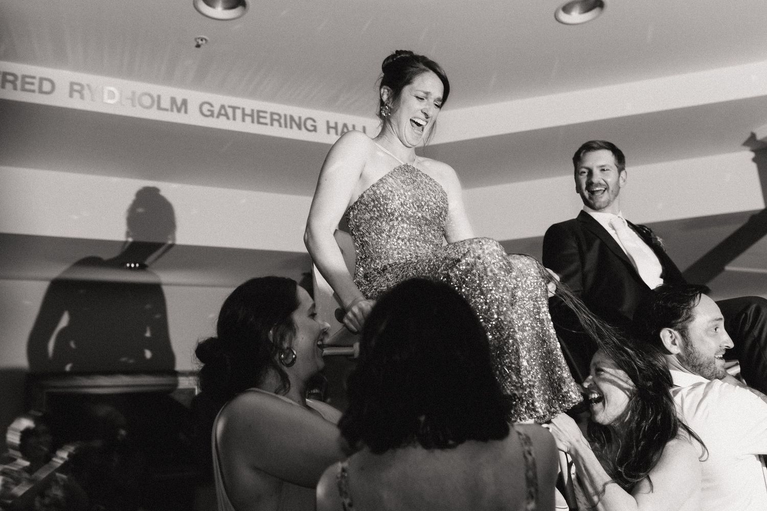 Wedding party guests lift a bride in a sparkling dress off the ground during a joyous celebration.