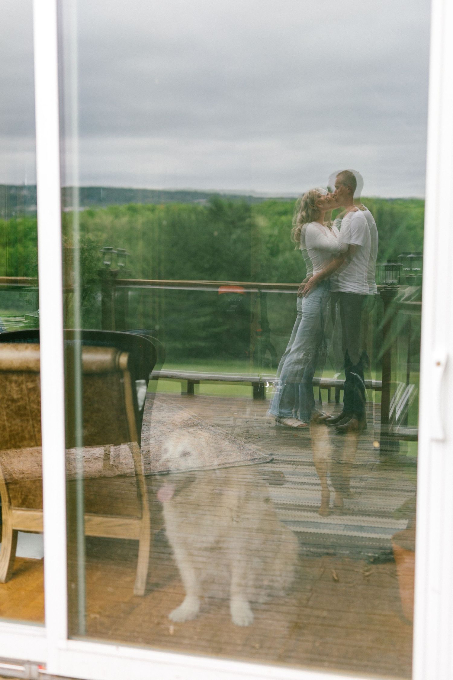 A couple embraces while standing on a balcony overlooking green countryside, with their reflection visible in a glass window.