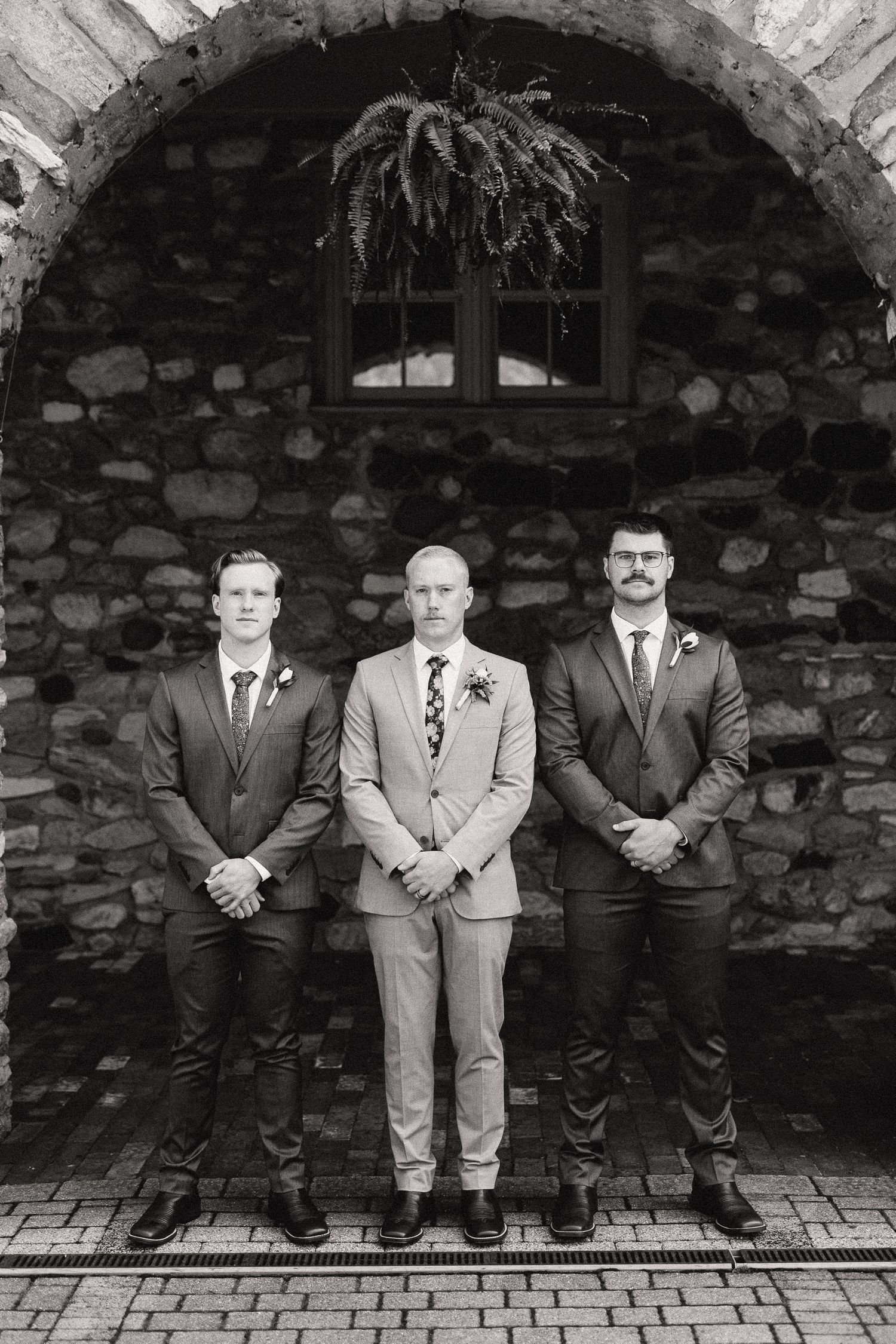 Groomsmen in formal suits pose against a rustic stone wall backdrop.