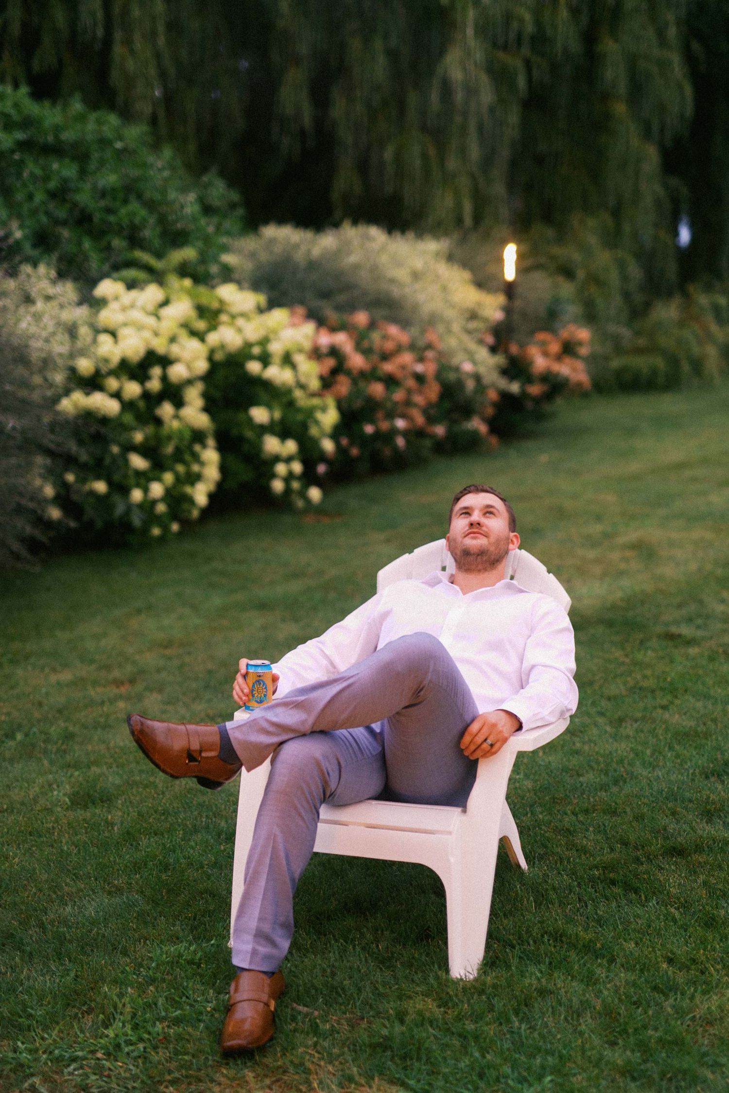 Someone relaxes in a white plastic Adirondack chair on a lawn with flowering shrubs and trees in the background at dusk.